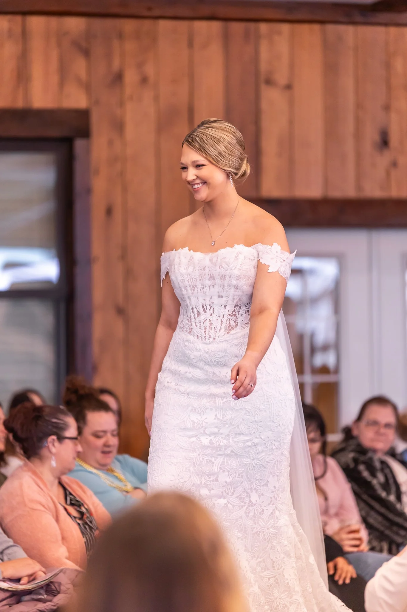 A woman in a white lace wedding dress smiling at a wedding ceremony with guests seated around her.