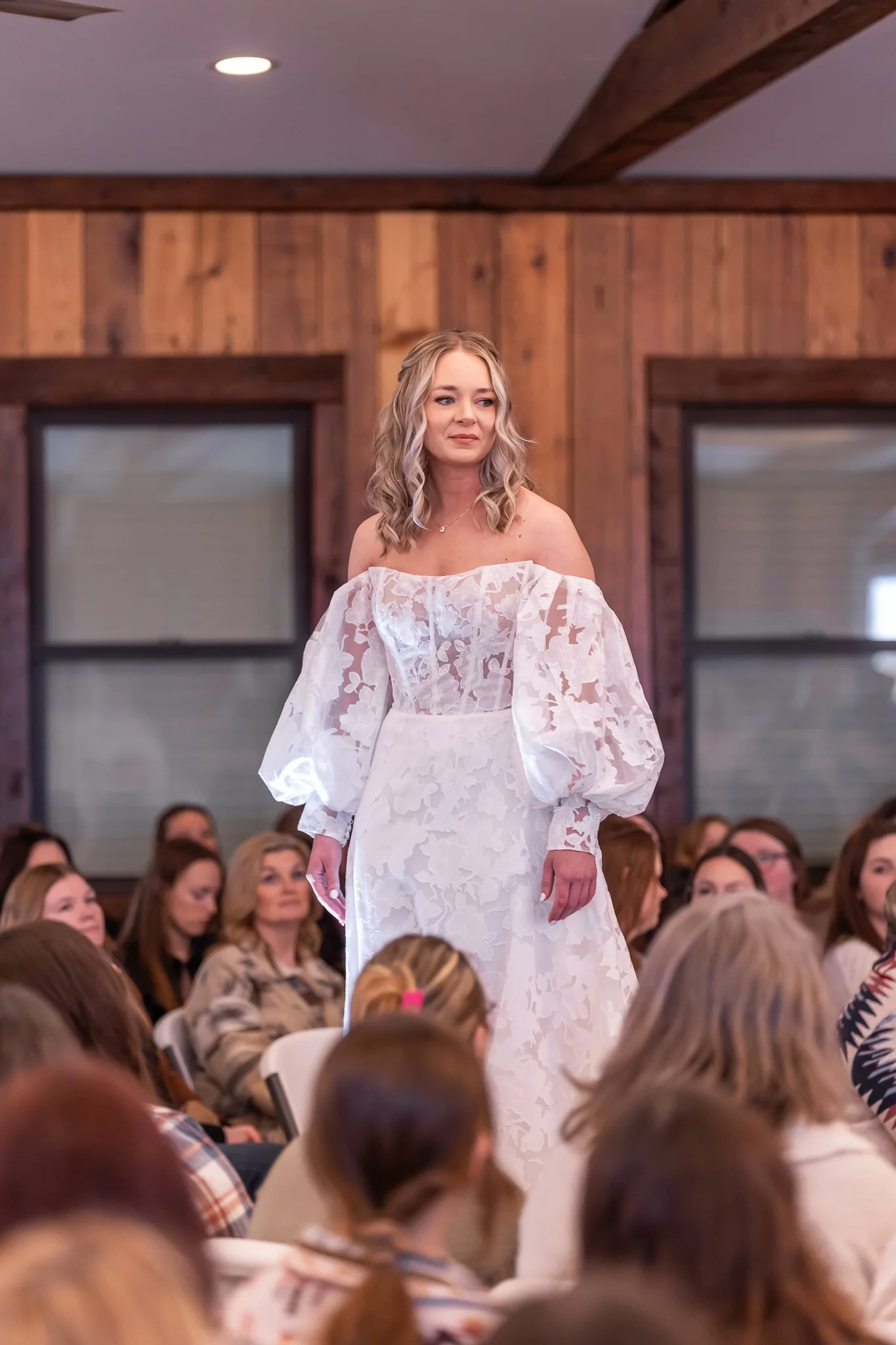 A woman with blonde hair wearing an off-the-shoulder white dress with lace detail walks in front of an audience in a room with wooden walls and large windows.