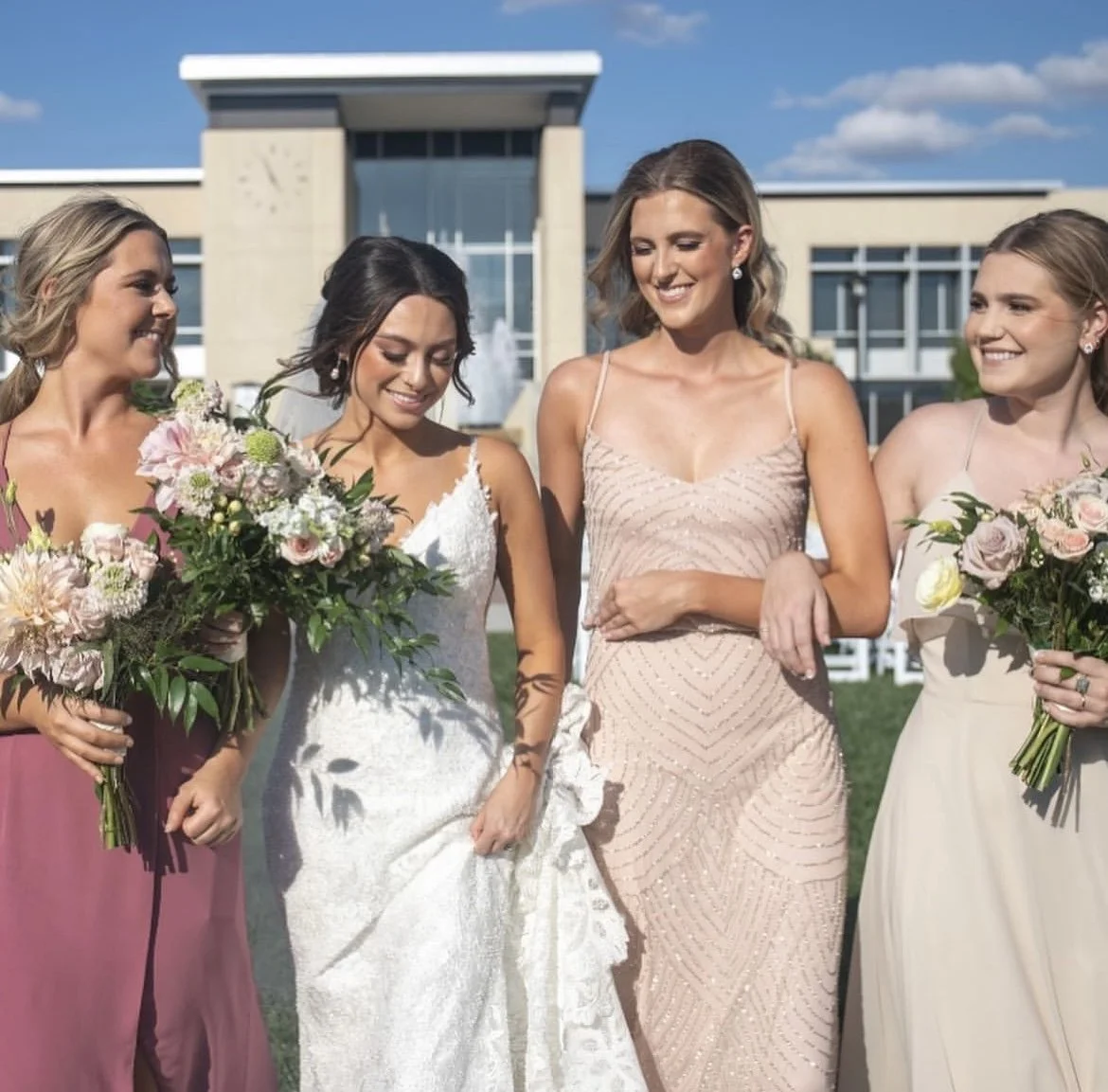 Four women in formal dresses standing outdoors, holding bouquets of flowers, smiling, with a modern building and blue sky in the background.