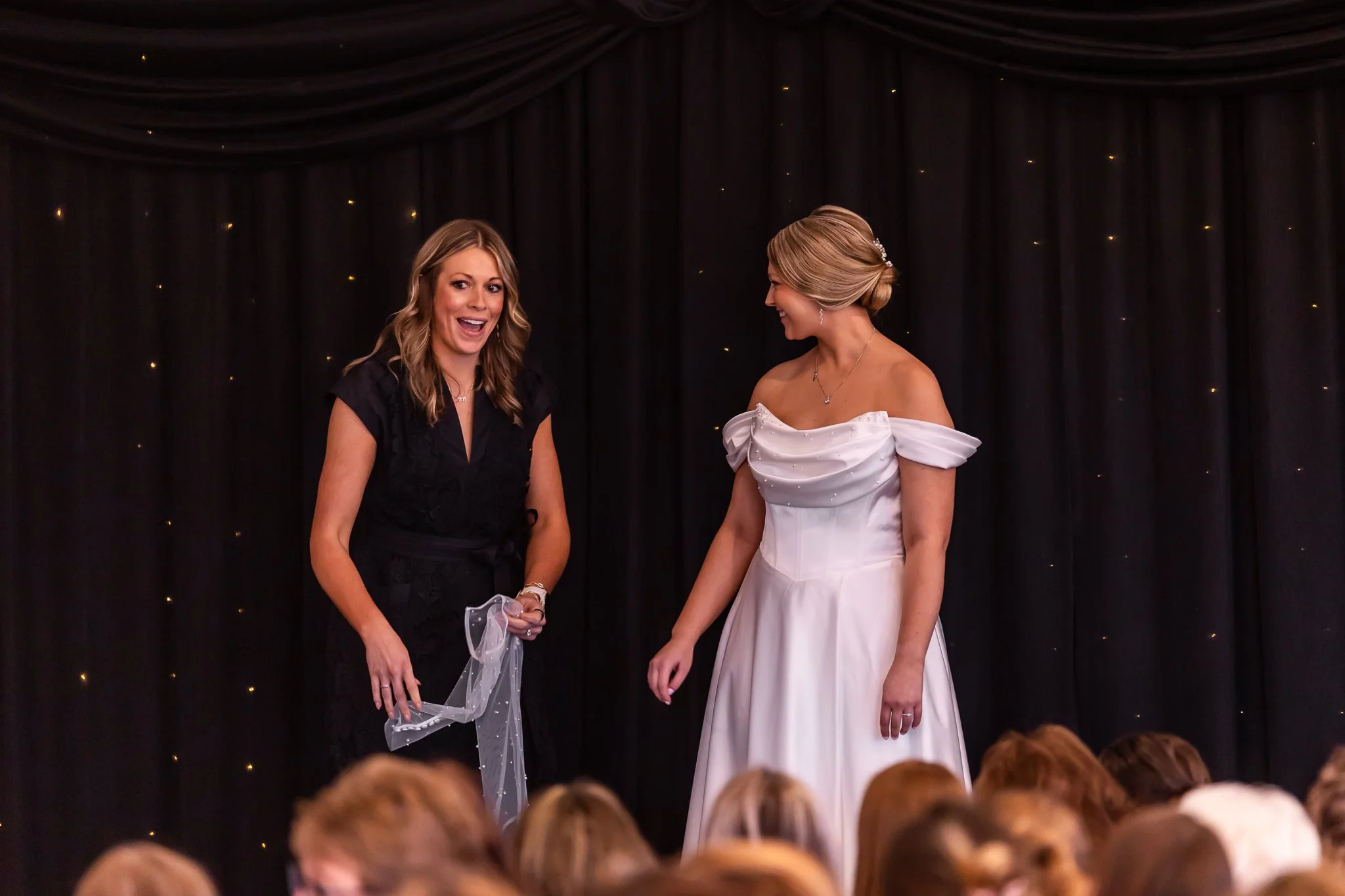 Two women standing on stage during a wedding reception, with a dark curtain backdrop and small lights, one in a black dress and the other in a white wedding gown, smiling and engaging in conversation.