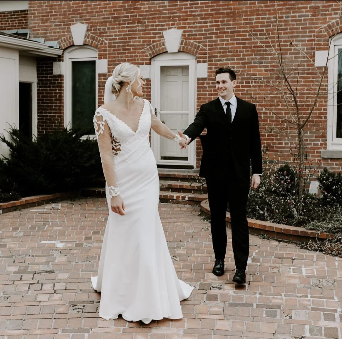 A bride and groom holding hands, standing outside a brick house, smiling at each other during their wedding.