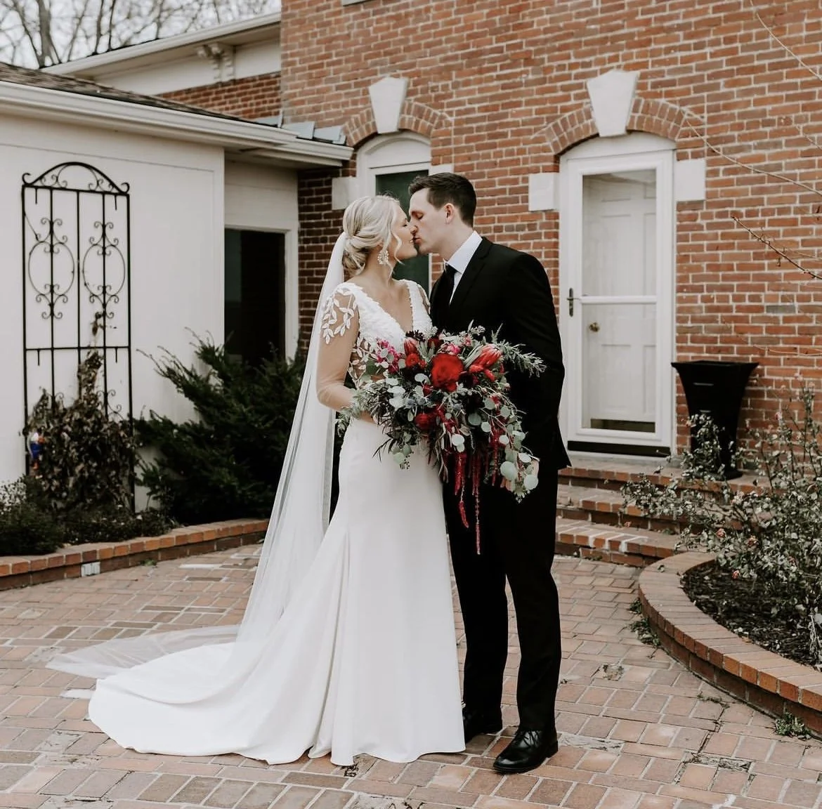 Bride and groom sharing a kiss outdoors, with the bride holding a bouquet of red flowers and greenery, in front of a brick building.