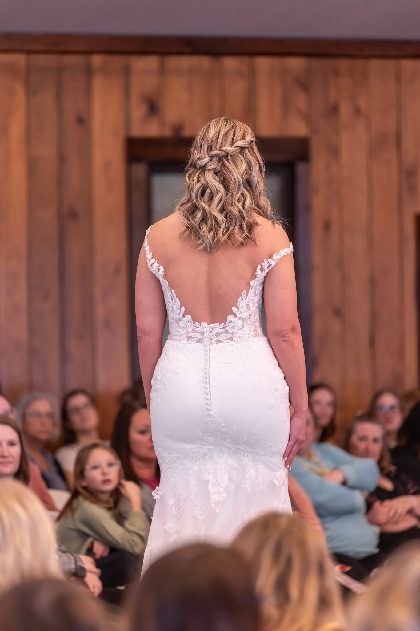 Bride in a white lace wedding gown with an open back, standing facing away in front of an audience.