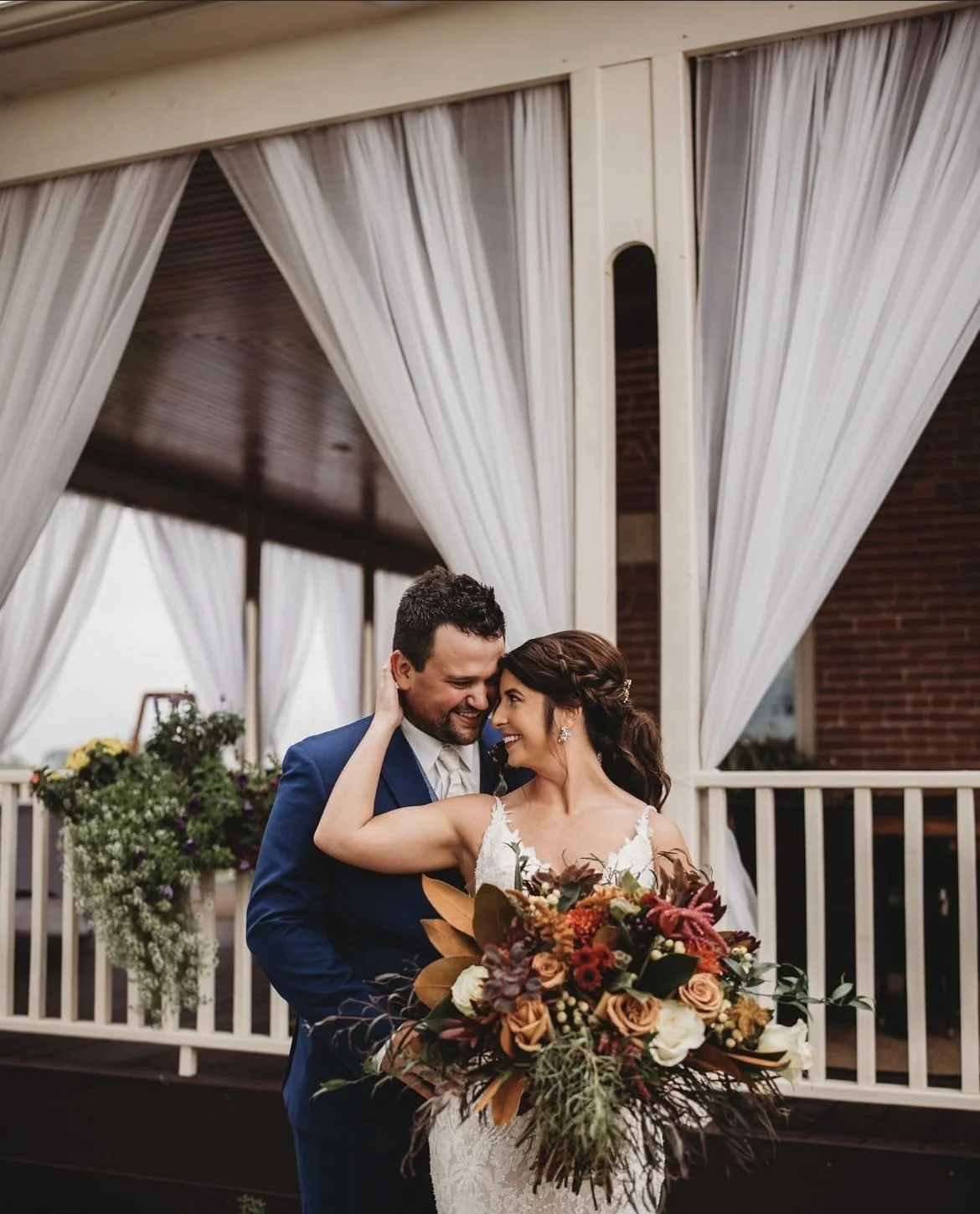 A bride and groom sharing a joyful moment, with the bride holding a large bouquet of flowers, standing on a porch with white curtains.