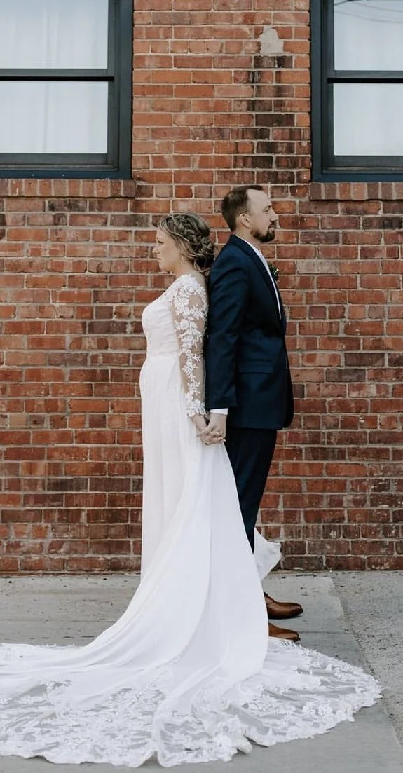 A bride in a white lace wedding dress and a groom in a dark suit, standing back-to-back and holding hands against a red brick wall.