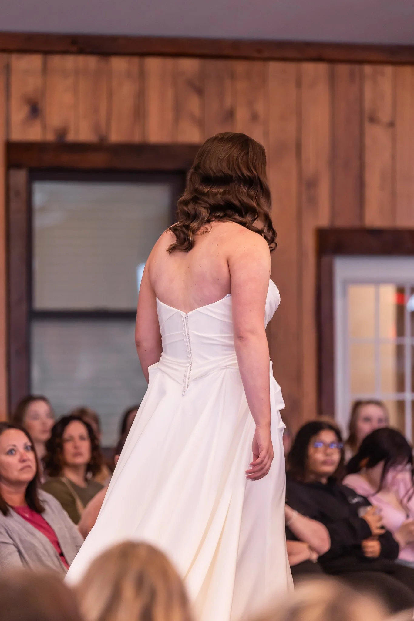 A woman in a white wedding dress stands with her back to the camera in front of an audience in a wood-paneled room.