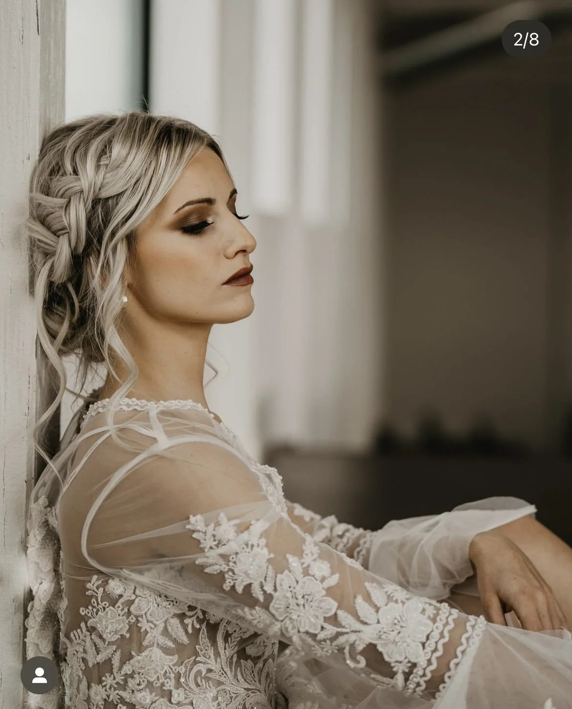 A woman with blonde hair styled in a braid and makeup in a white lace dress sitting against a wall.