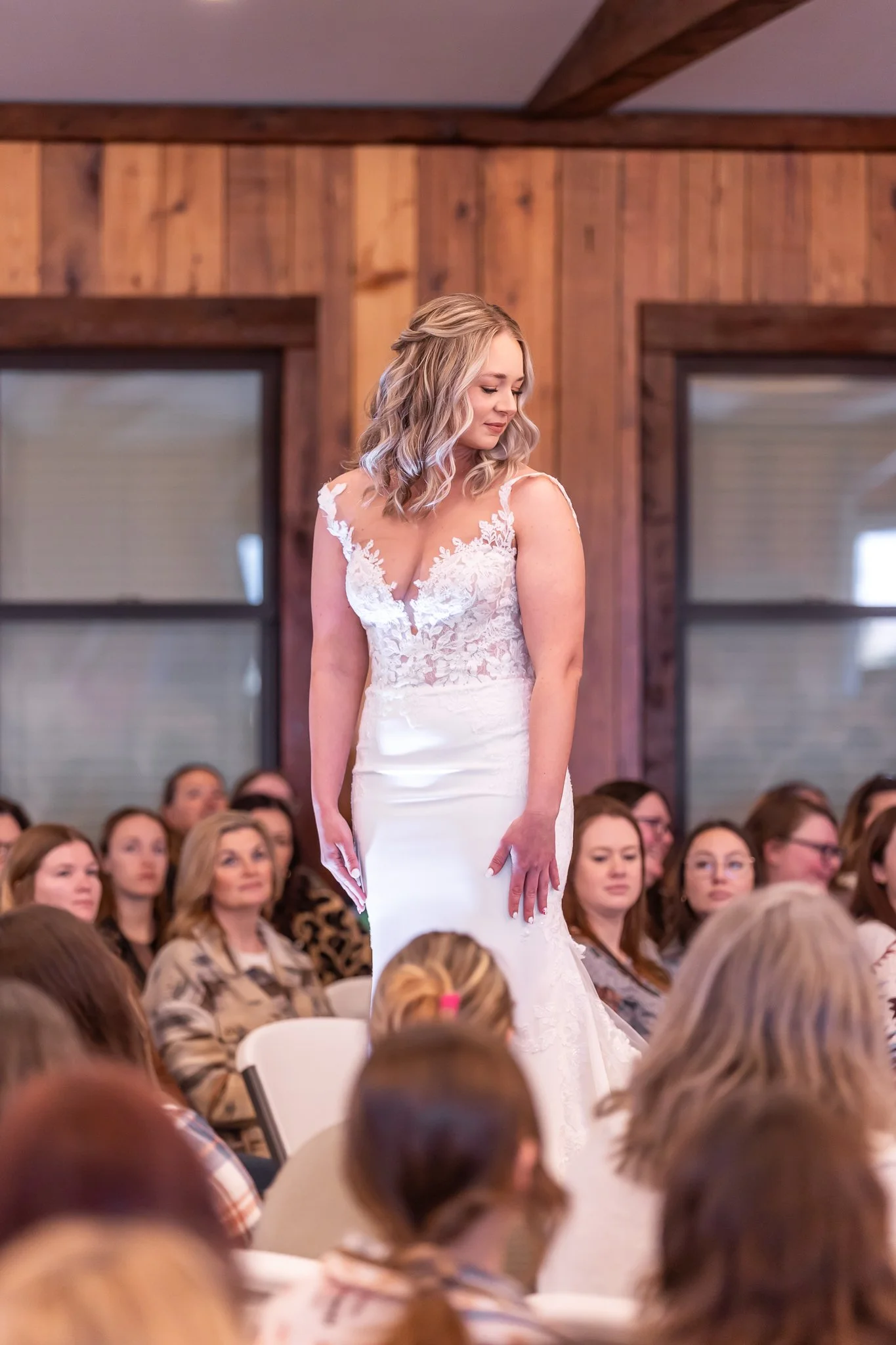 A bride in a white lace wedding gown standing among seated guests in a rustic venue with wood-paneled walls and large windows.