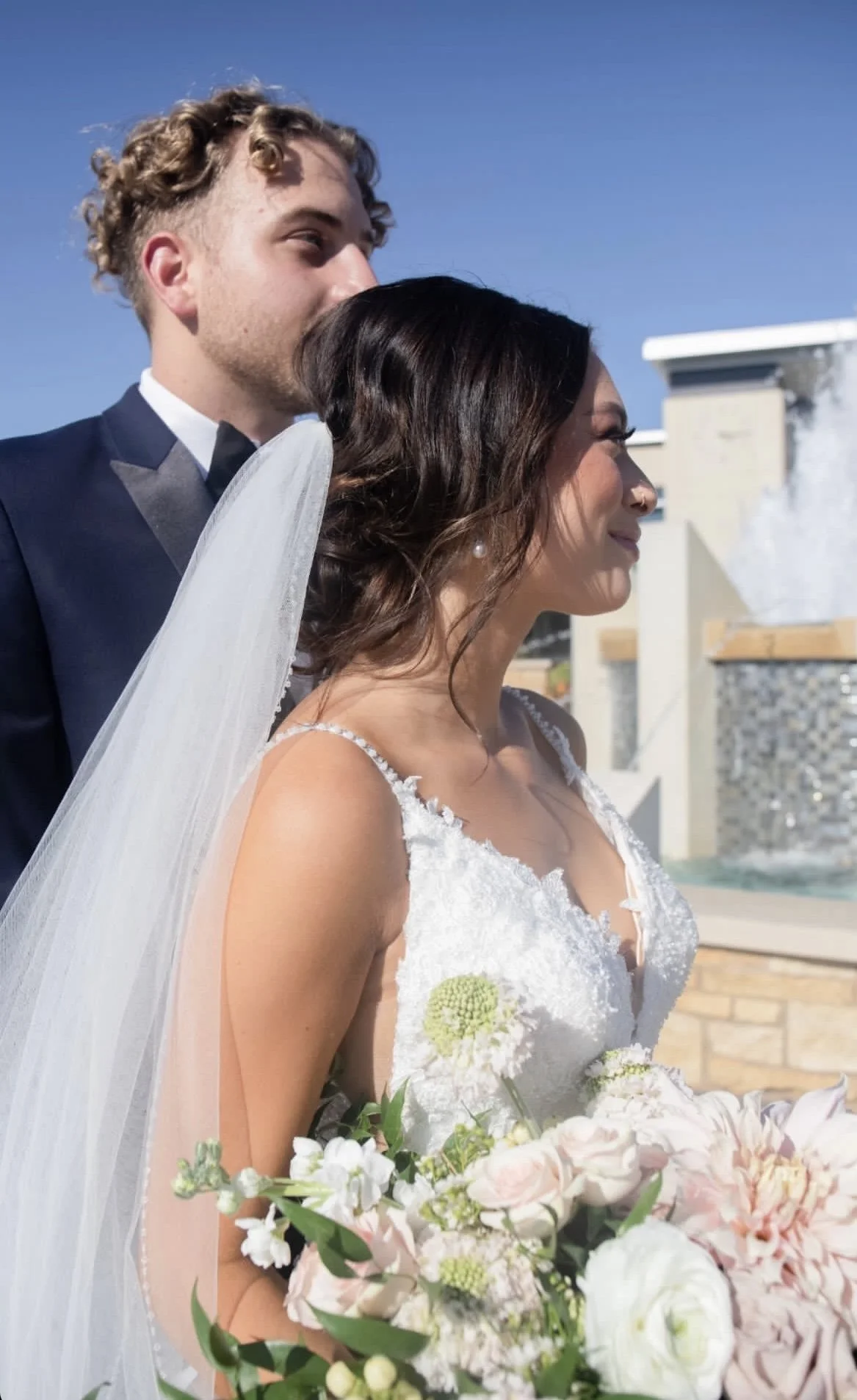 A bride and groom embrace outdoors, with a fountain and blue sky in the background after their wedding.