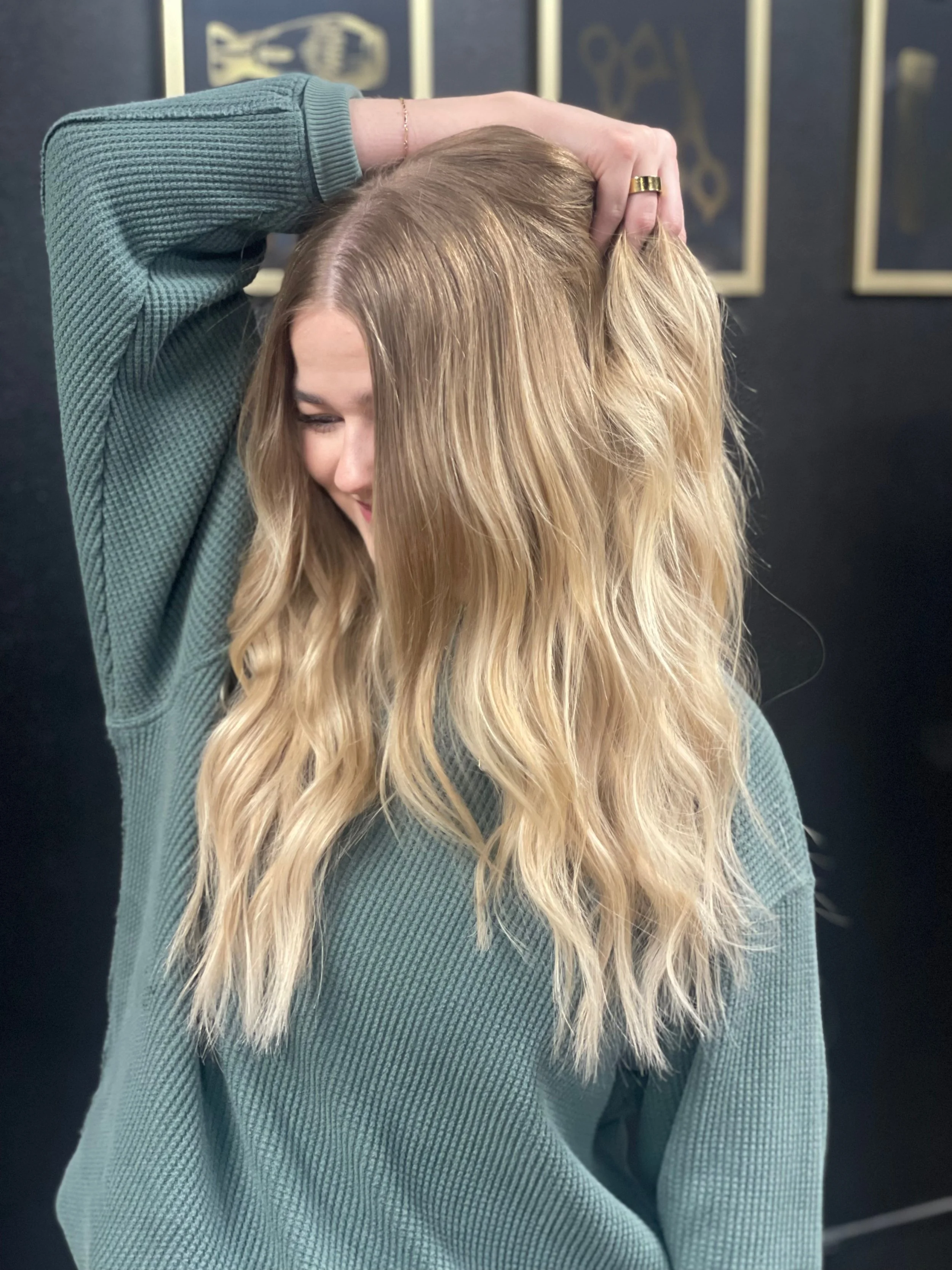 A woman with long, wavy blonde hair touching her hair and smiling, wearing a green textured long sleeve top, standing against a dark background with framed pictures.