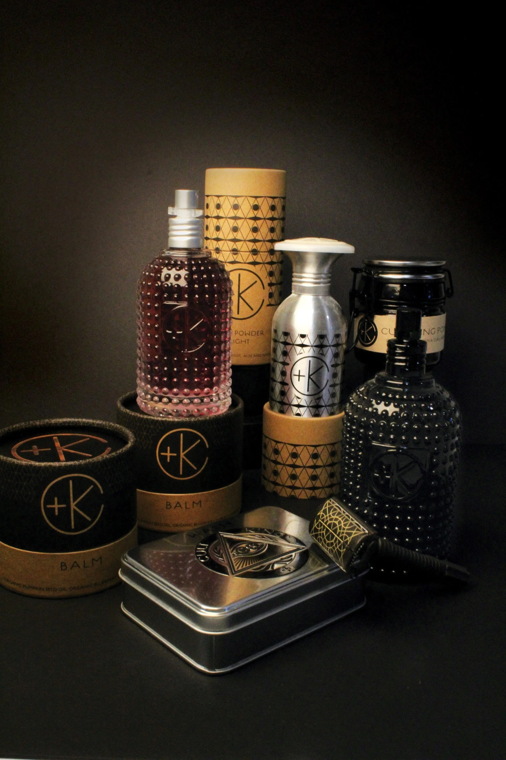 Collection of skincare and beauty products including bottles, jars, a metal tin, and a jade roller, all with a matching black and gold design.