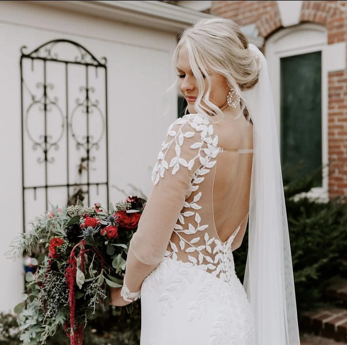 Bride in a wedding dress with open back and floral lace details holding a bouquet of red and pink flowers outdoors.
