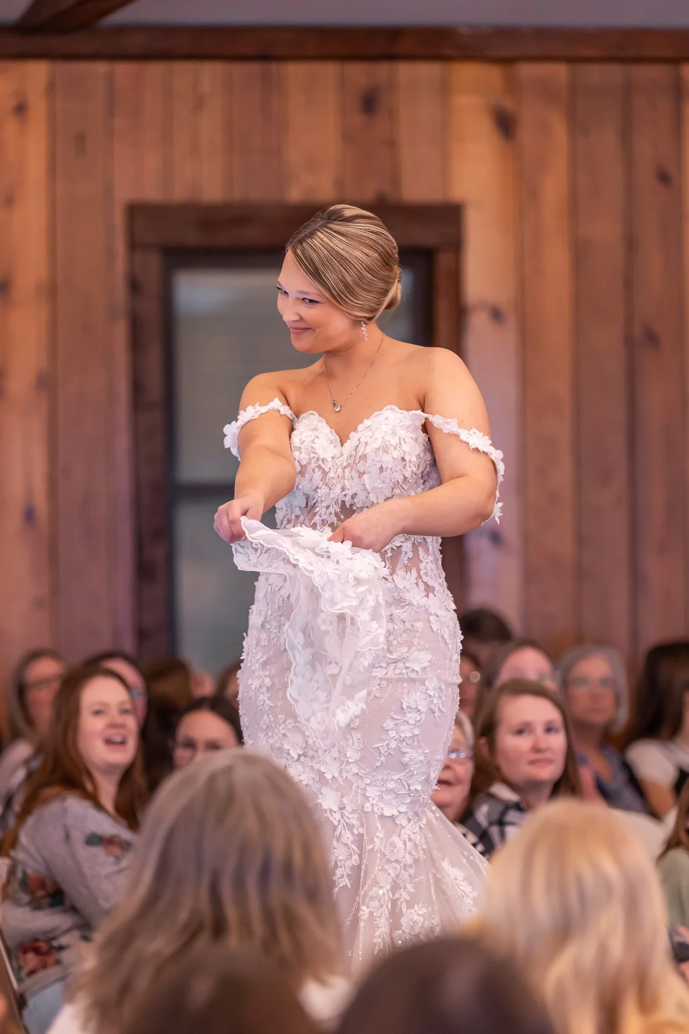 A woman in a wedding dress is holding and examining her veil during a wedding ceremony or reception, with a gathered crowd of guests watching and smiling.