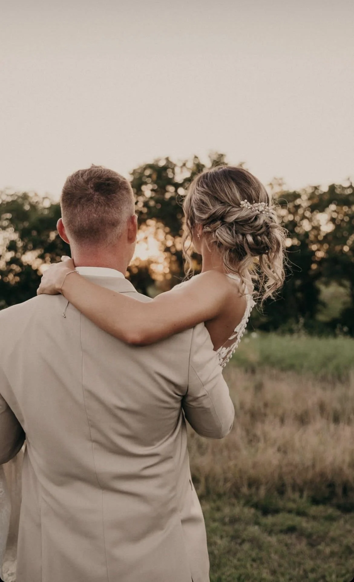 A newlywed couple, with the groom holding the bride, walks through a grassy field at sunset. The bride has a floral hair accessory, and the background features trees and a warm sky.