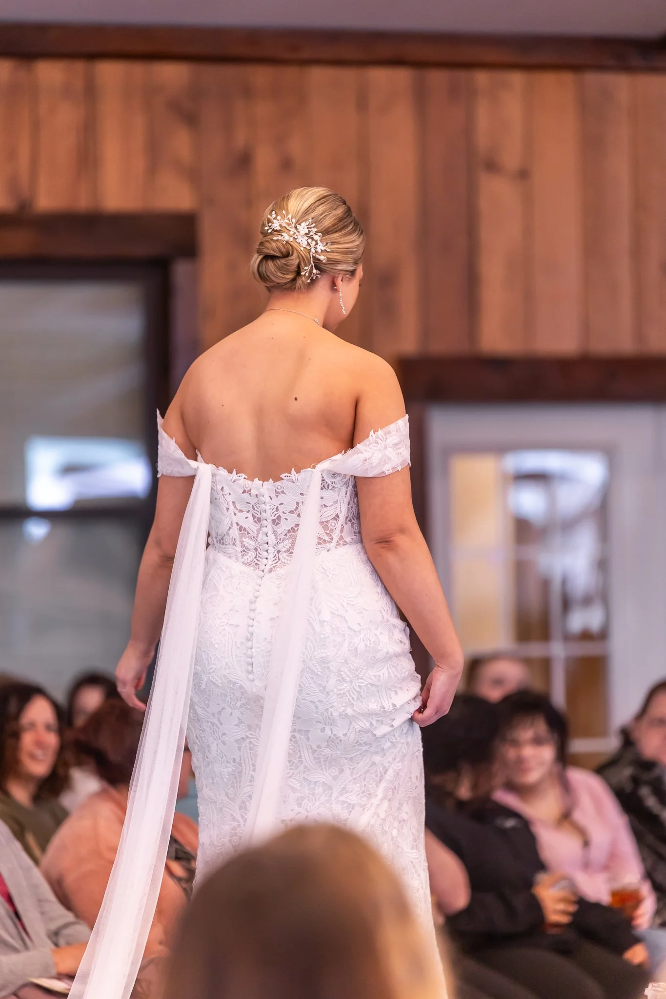 A woman in a white lace wedding dress with off-the-shoulder sleeves, standing with her back to the camera, showcasing her elegant updo hairstyle with a decorative hairpiece, in a room with wooden walls and an audience seated and watching her.