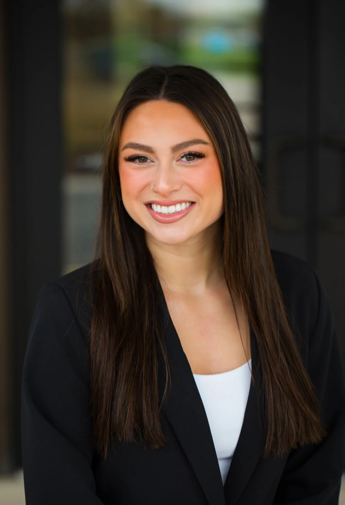 A young woman with long brown hair, smiling, wearing a black blazer and a white top, standing in front of a blurred background.