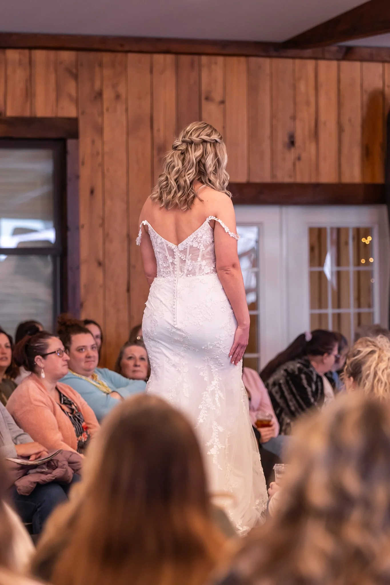 A woman in a white wedding dress standing in front of seated guests at a wedding reception, with her back to the camera, in a room with wooden walls and a glass door.