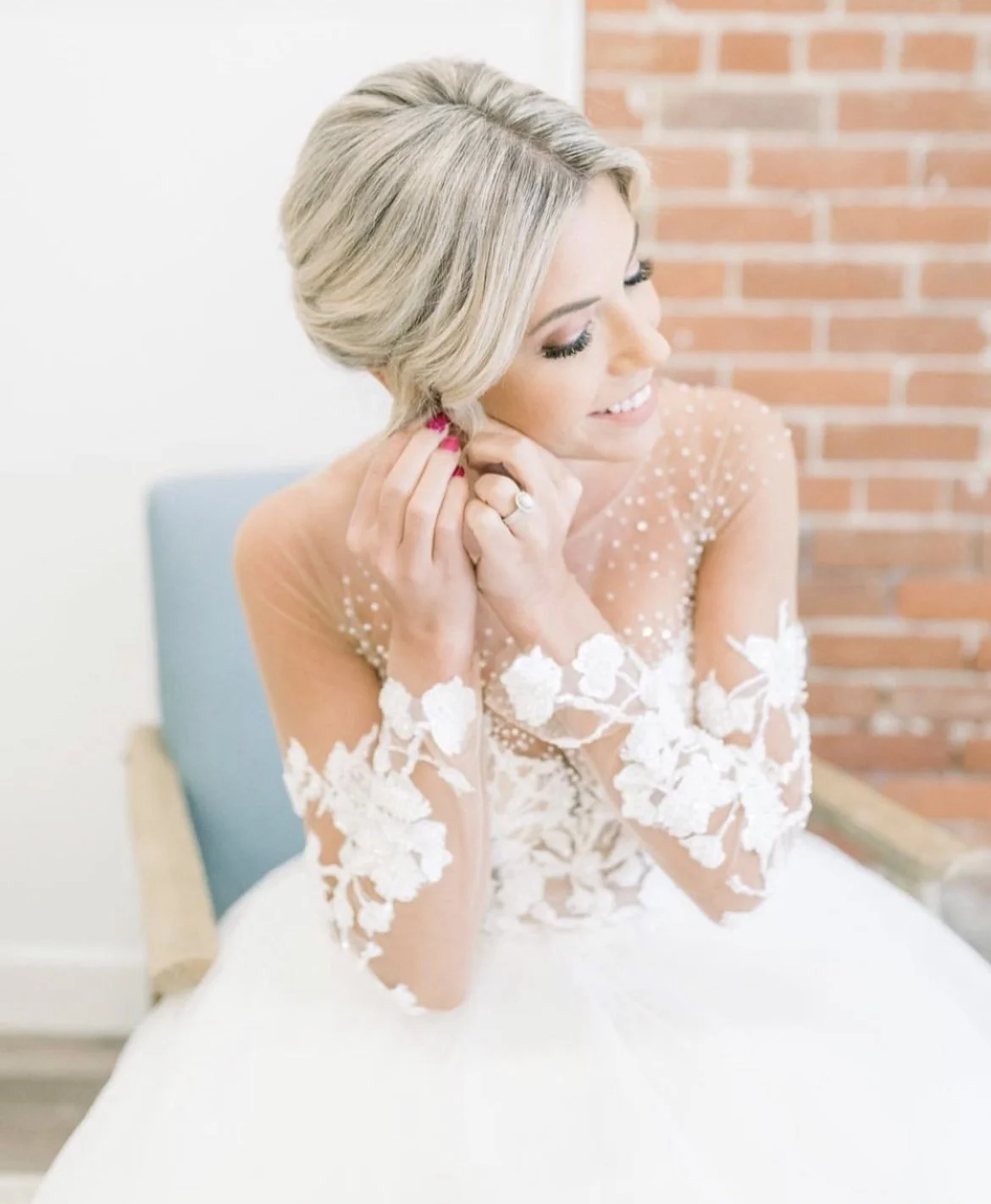 Bride getting ready, sitting on a bench, putting on earrings, in a white wedding dress with floral lace sleeves, in front of a brick wall.