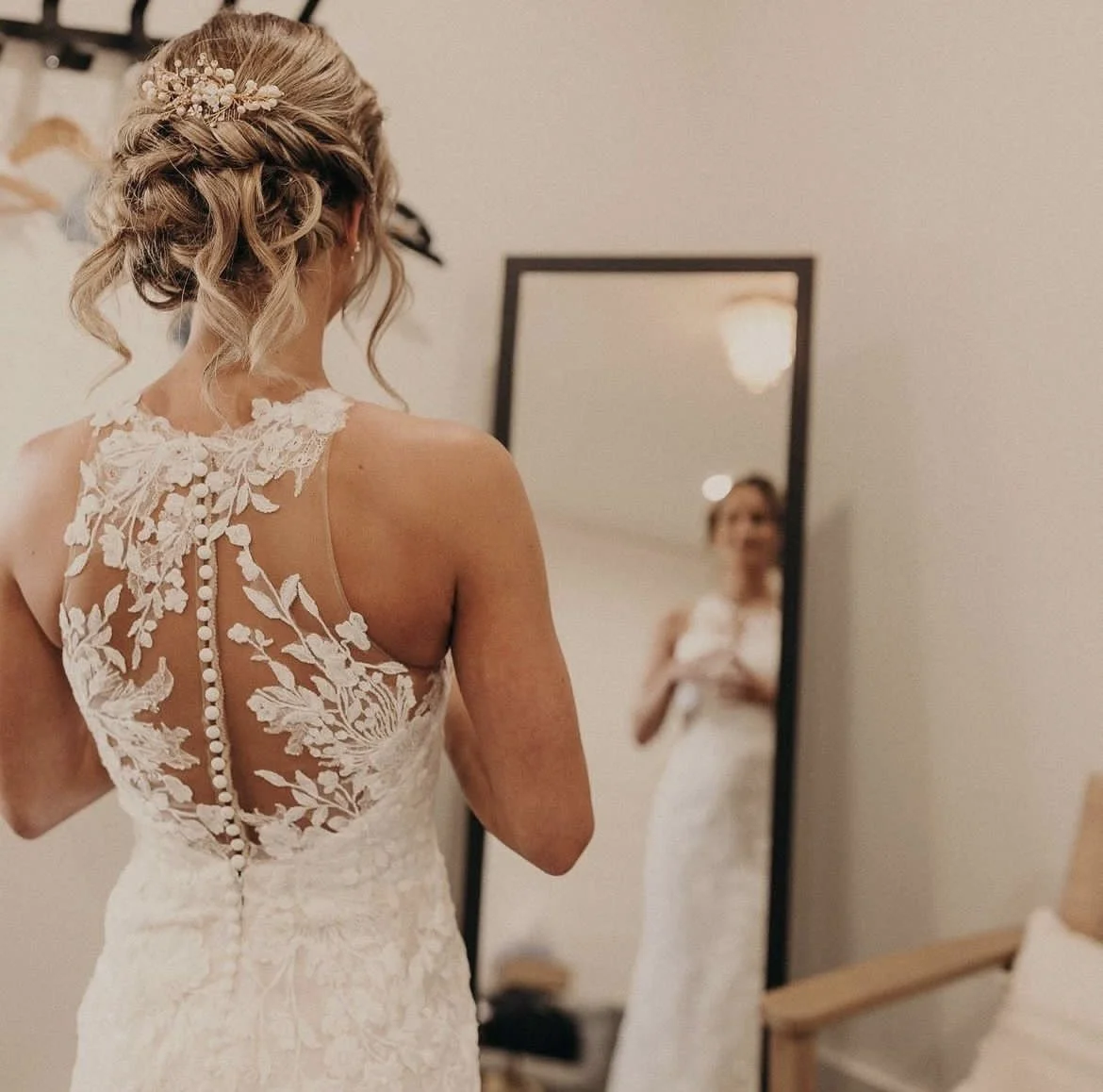 A bride in a white lace wedding dress with floral embroidery looks at herself in a mirror before her wedding.
