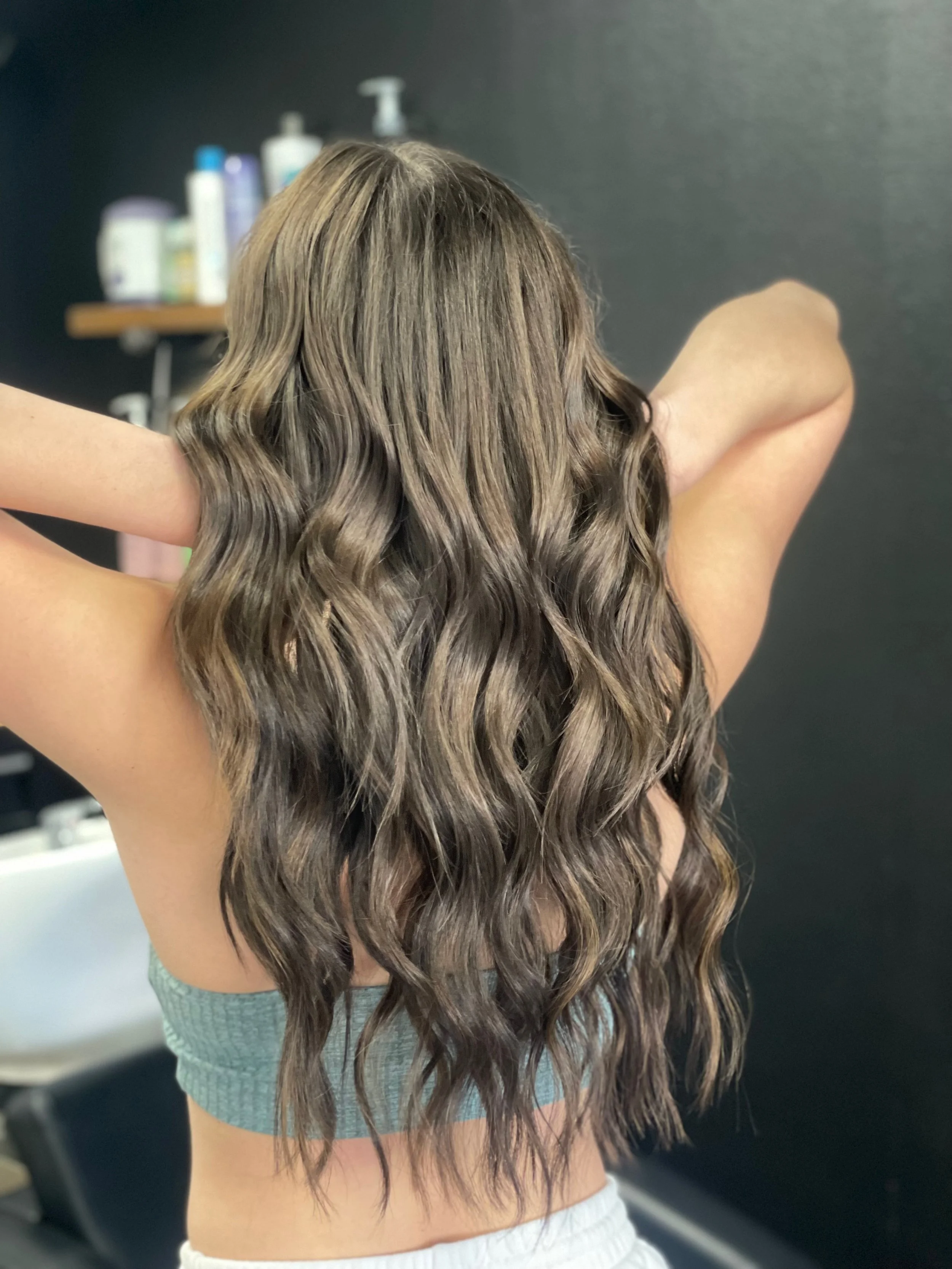 Back view of a woman with long, wavy brown hair in a salon or bathroom setting.