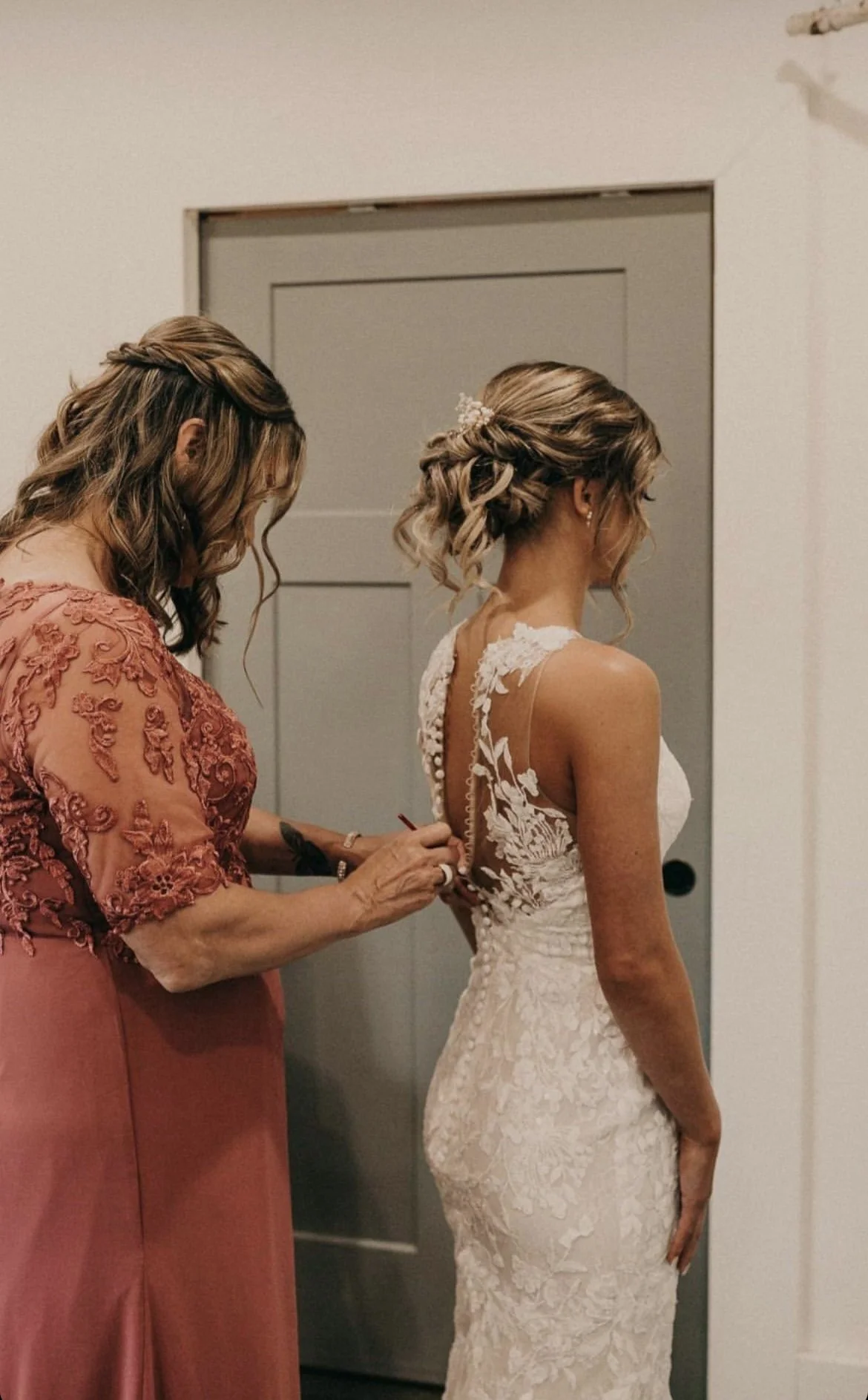A bride standing in a wedding dress while a woman helps her zip up the back of the dress.