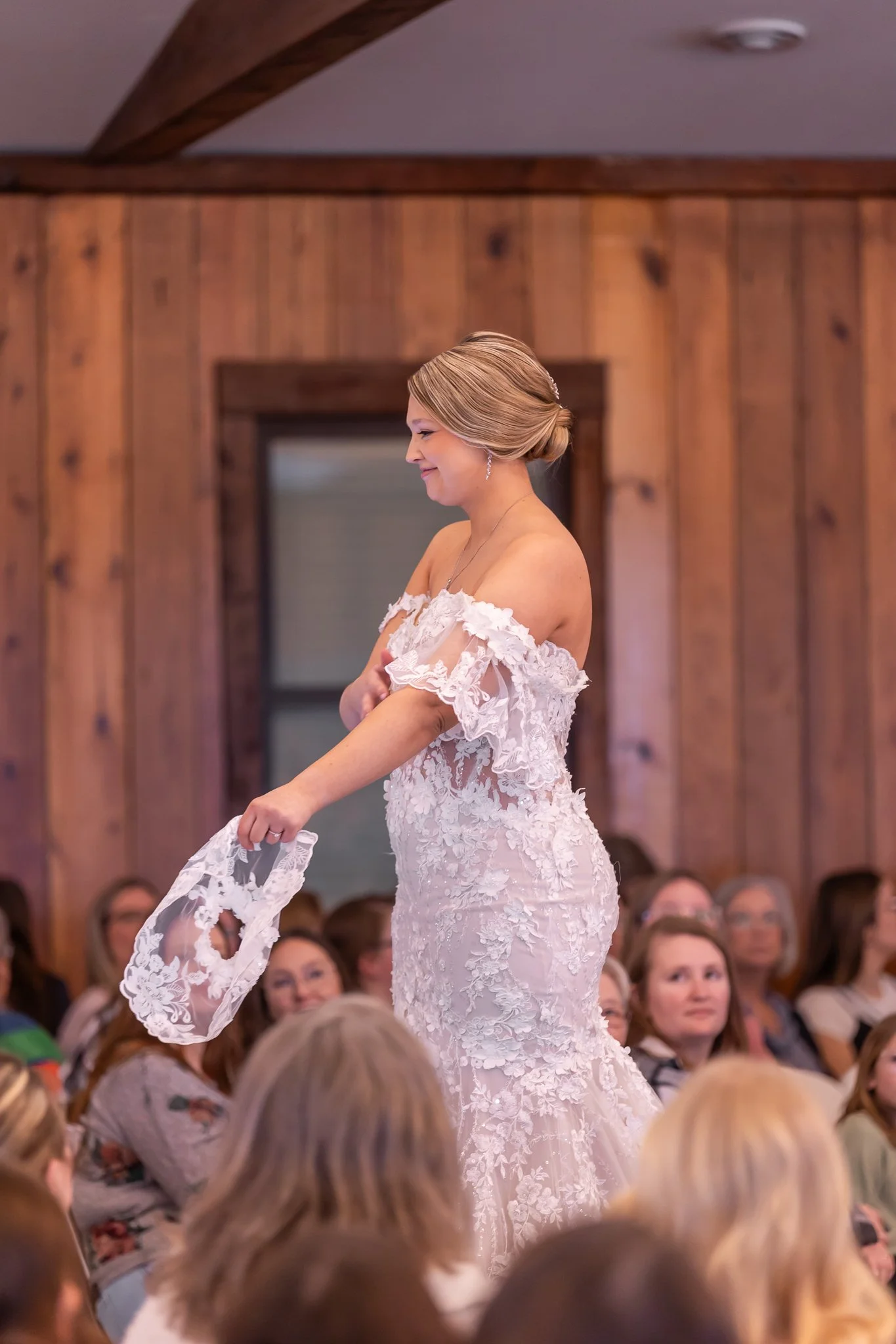 A bride in a white lace wedding dress holding her bouquet, smiling at her wedding reception with guests seated around her in a wood-paneled room.