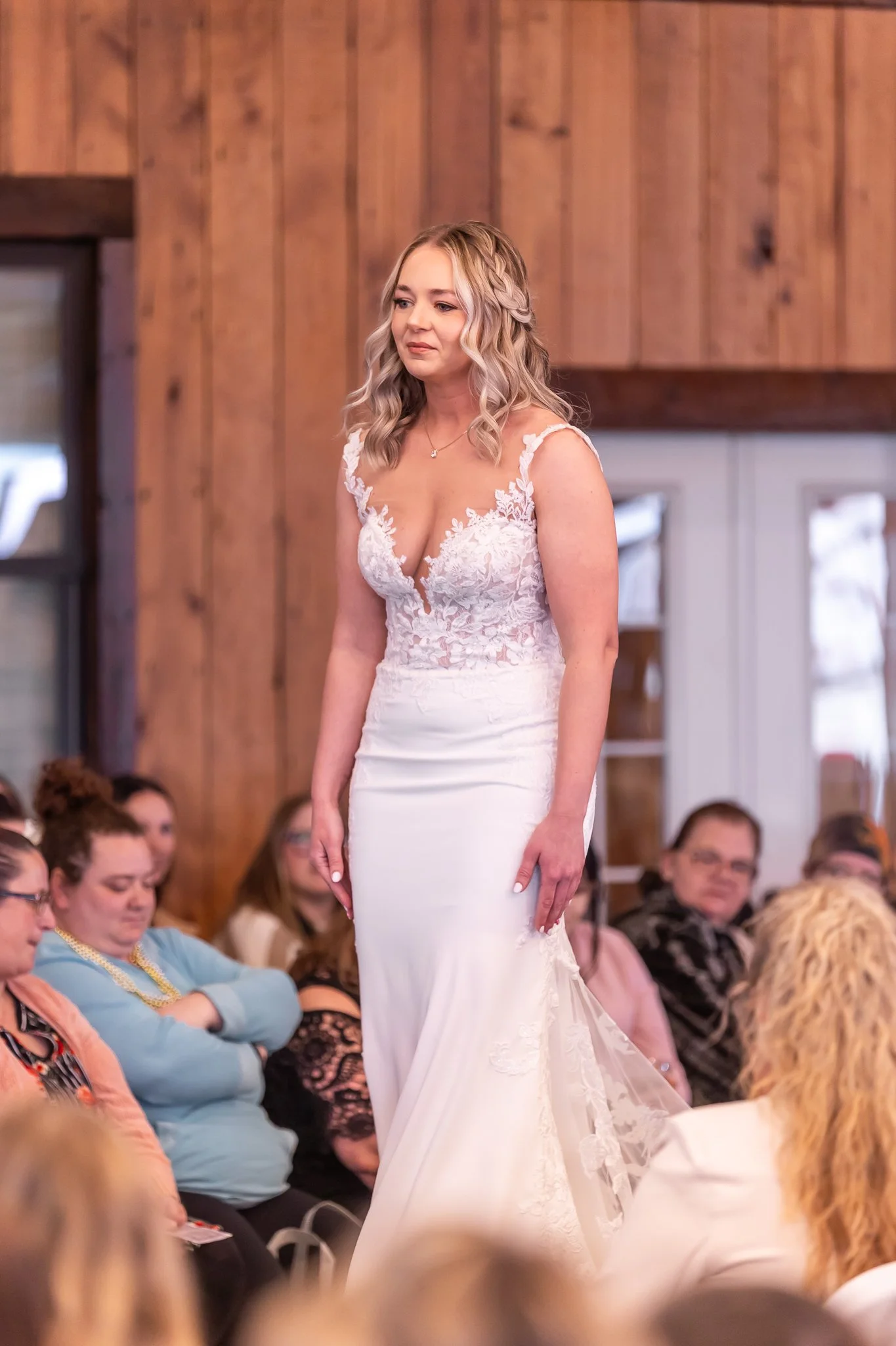 A woman in a white wedding dress walking down the aisle with guests seated on either side, in a wooden interior setting.