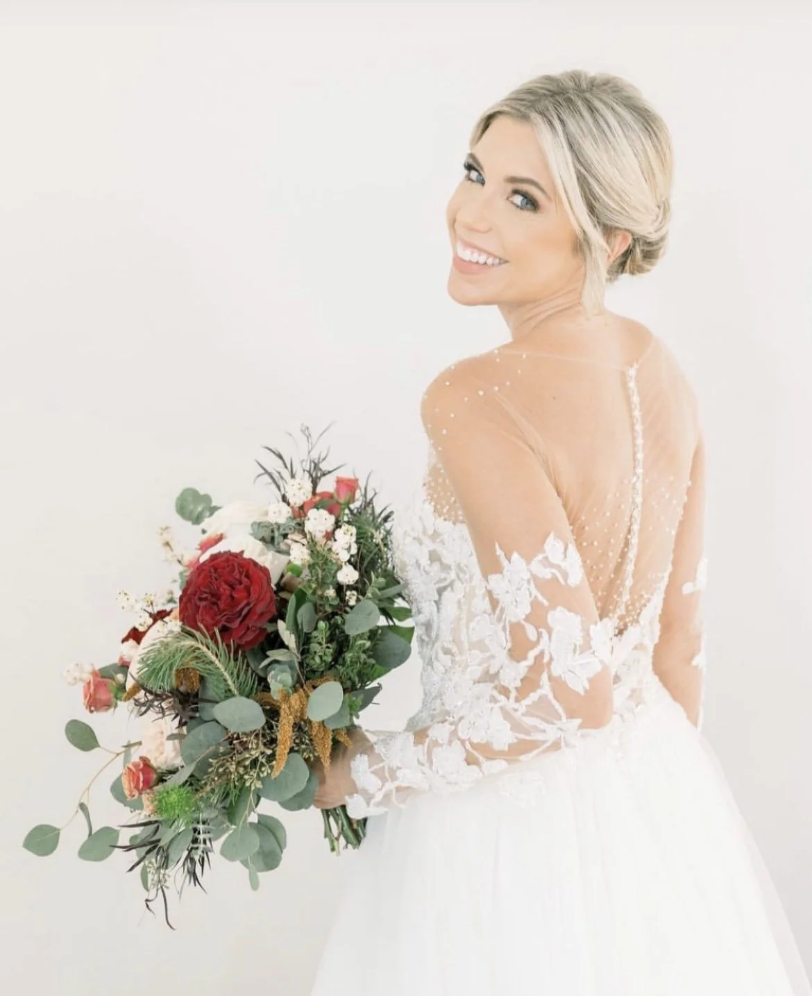 A smiling bride with blonde hair in a wedding dress holding a bouquet of red, white, and green flowers against a white background.