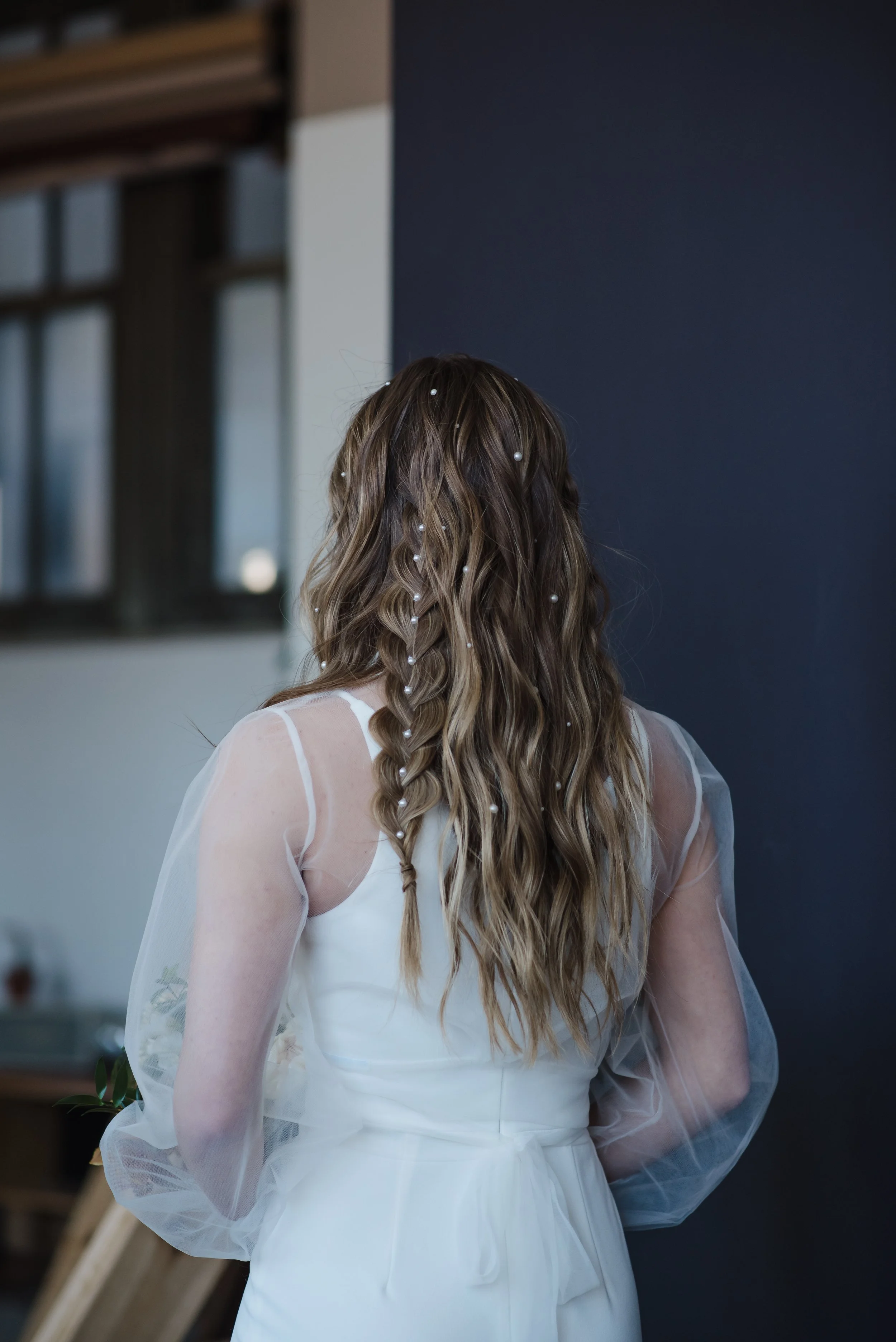 A woman with long, wavy hair decorated with pearls, wearing a white dress with sheer puffed sleeves, stands indoors facing away from the camera.