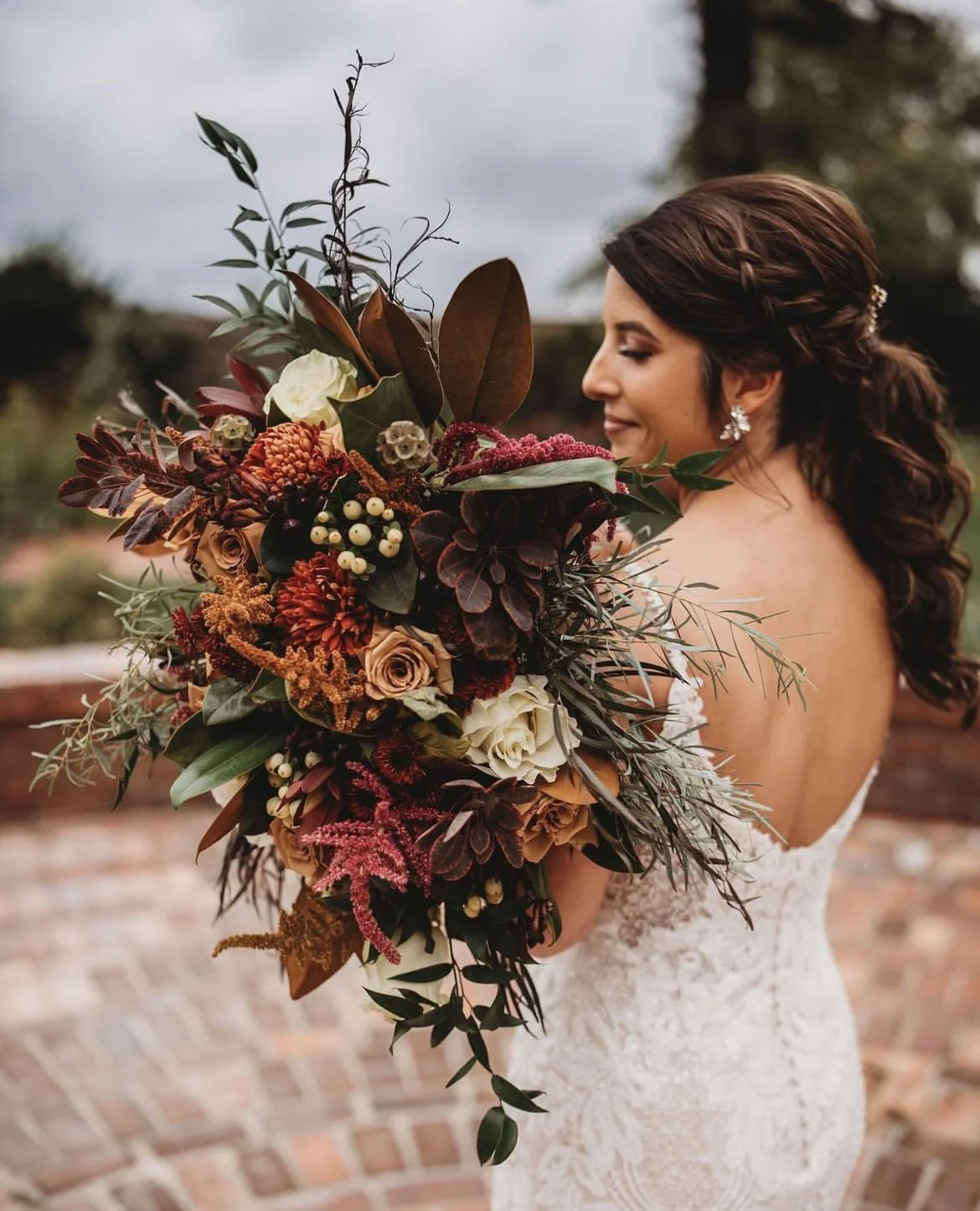 Bride holding a large bouquet of red, orange, white, and brown flowers with greenery, wearing a strapless lace wedding dress, outdoors with a brick pathway.