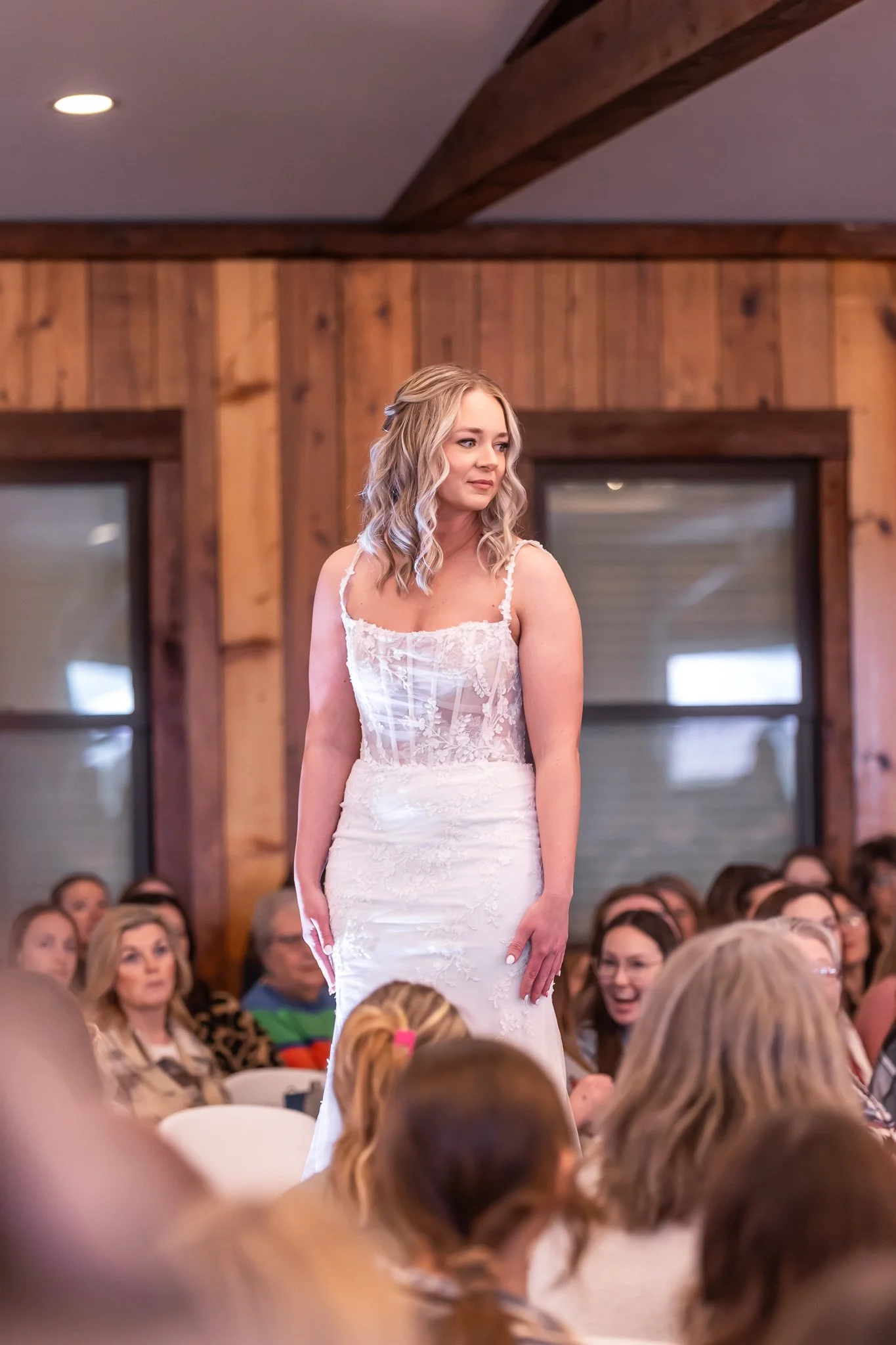 A bride in a white lace wedding gown standing indoors, surrounded by seated guests, wooden walls, and large windows in the background.