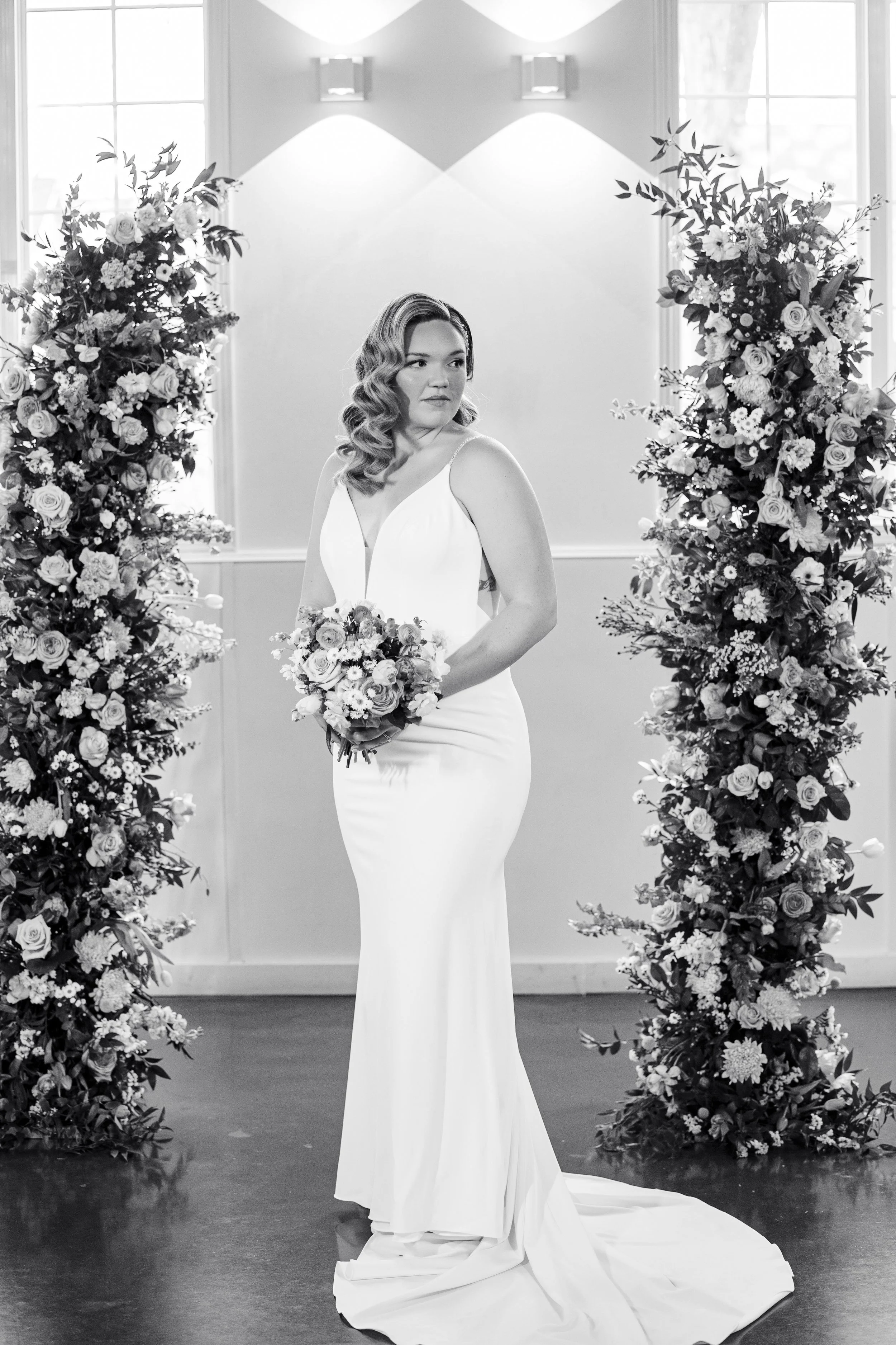 A woman in a white wedding dress holding a bouquet of flowers stands between two large floral arrangements in an indoor setting with windows and wall lights.