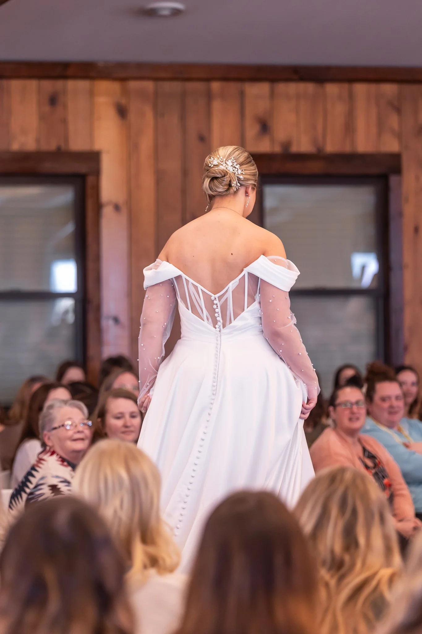 A bride in a wedding gown with sheer, beaded long sleeves and an open back, standing in front of an audience in a room with wooden paneling, as she prepares to walk down the aisle.