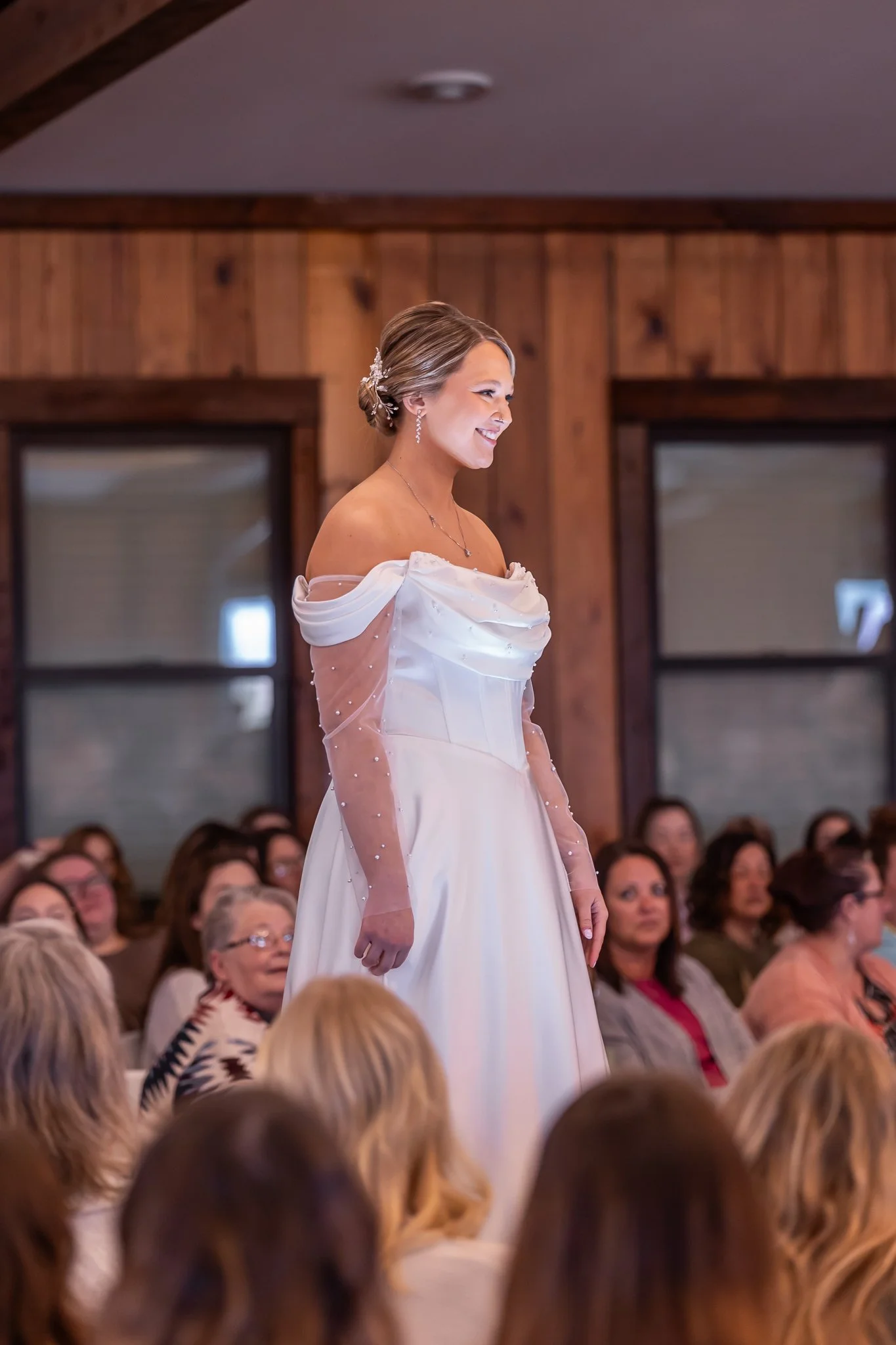A woman in a wedding dress smiles while standing in front of seated guests in a wooden room.