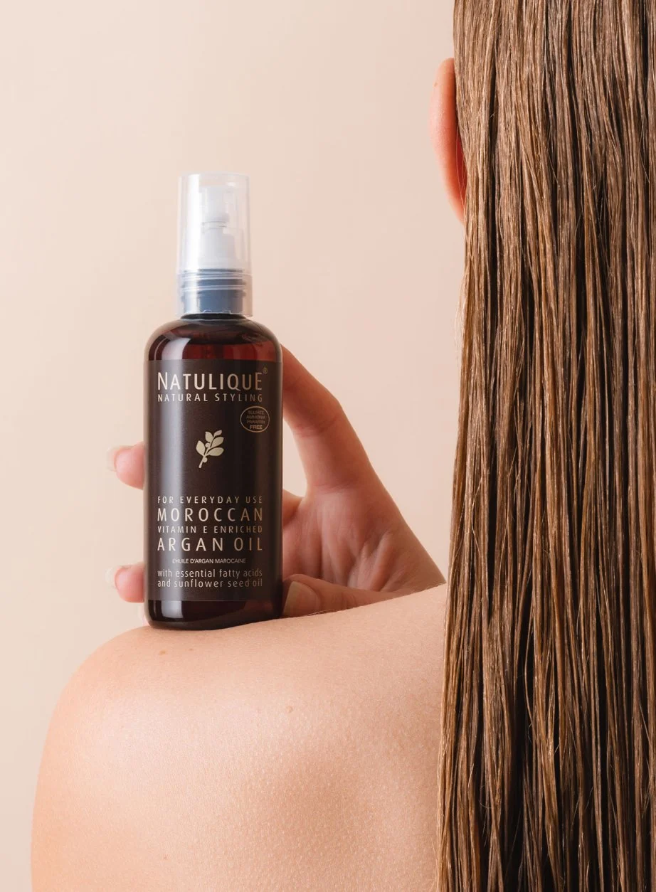 A woman with long wet brown hair holding a bottle of Natulique Moroccan argan oil for everyday use near her shoulder, against a plain beige background.