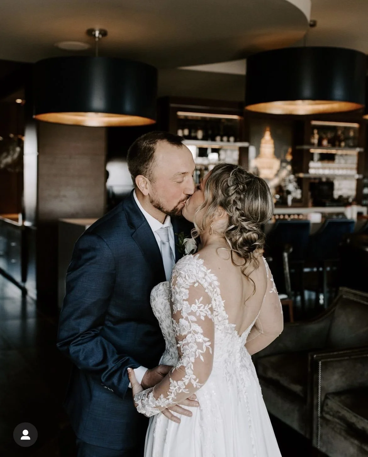 A newlywed couple, dressed in wedding attire, sharing a kiss indoors with a bar in the background.