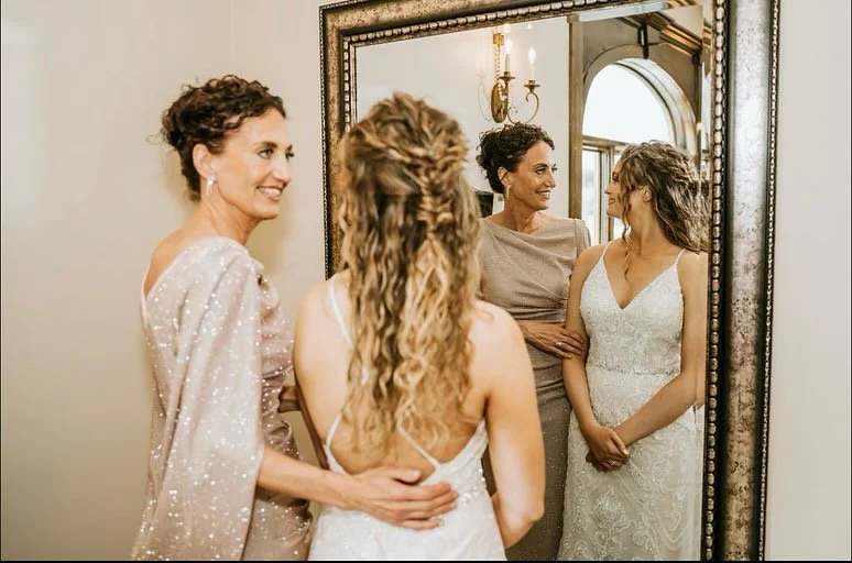 Three women looking at their reflection in a mirror, two of them in wedding dresses and one in a bronze-colored dress, in a room with elegant lighting and decor.