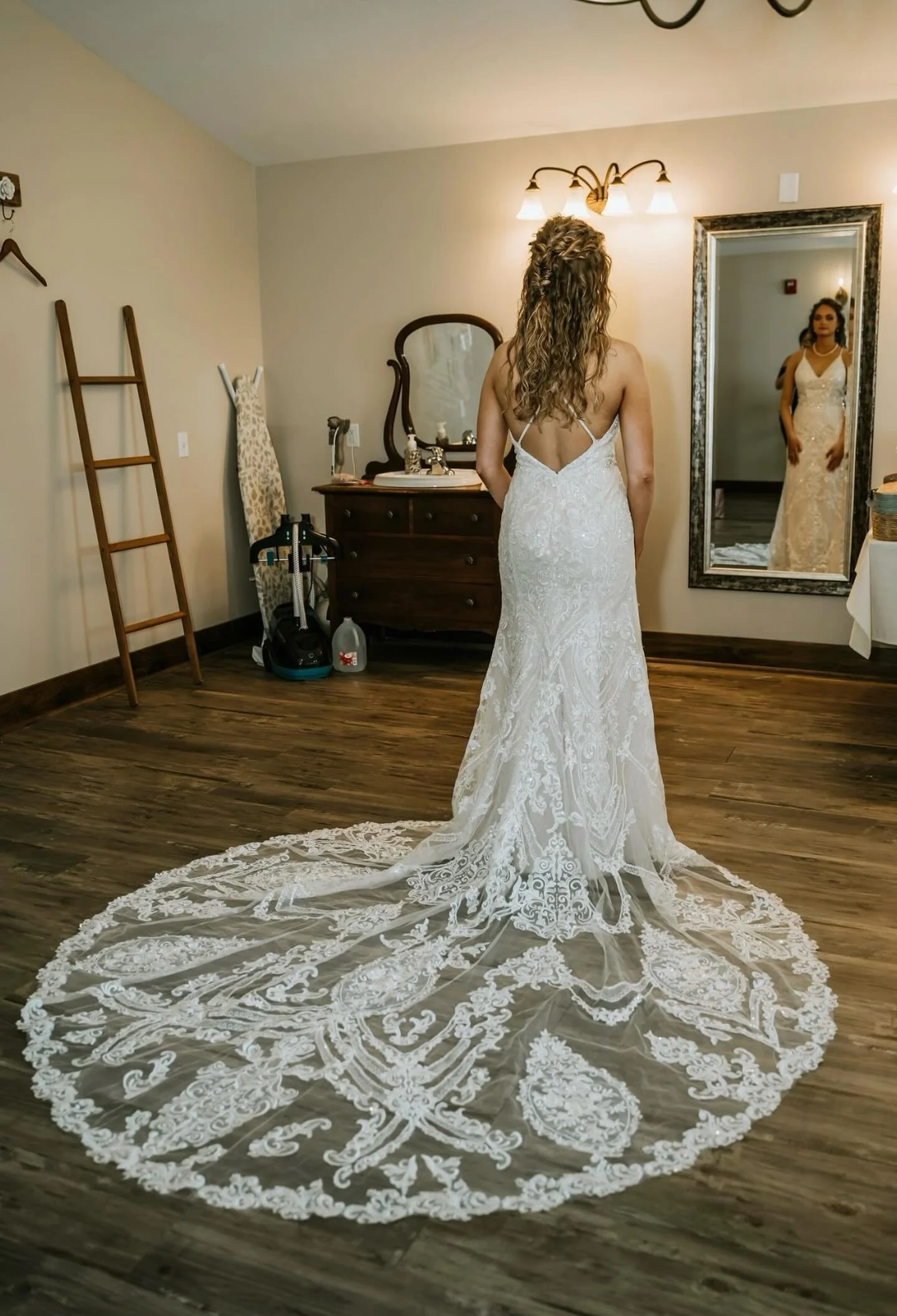 A bride in a white lace wedding gown with a long train standing in front of a mirror, looking at her reflection.