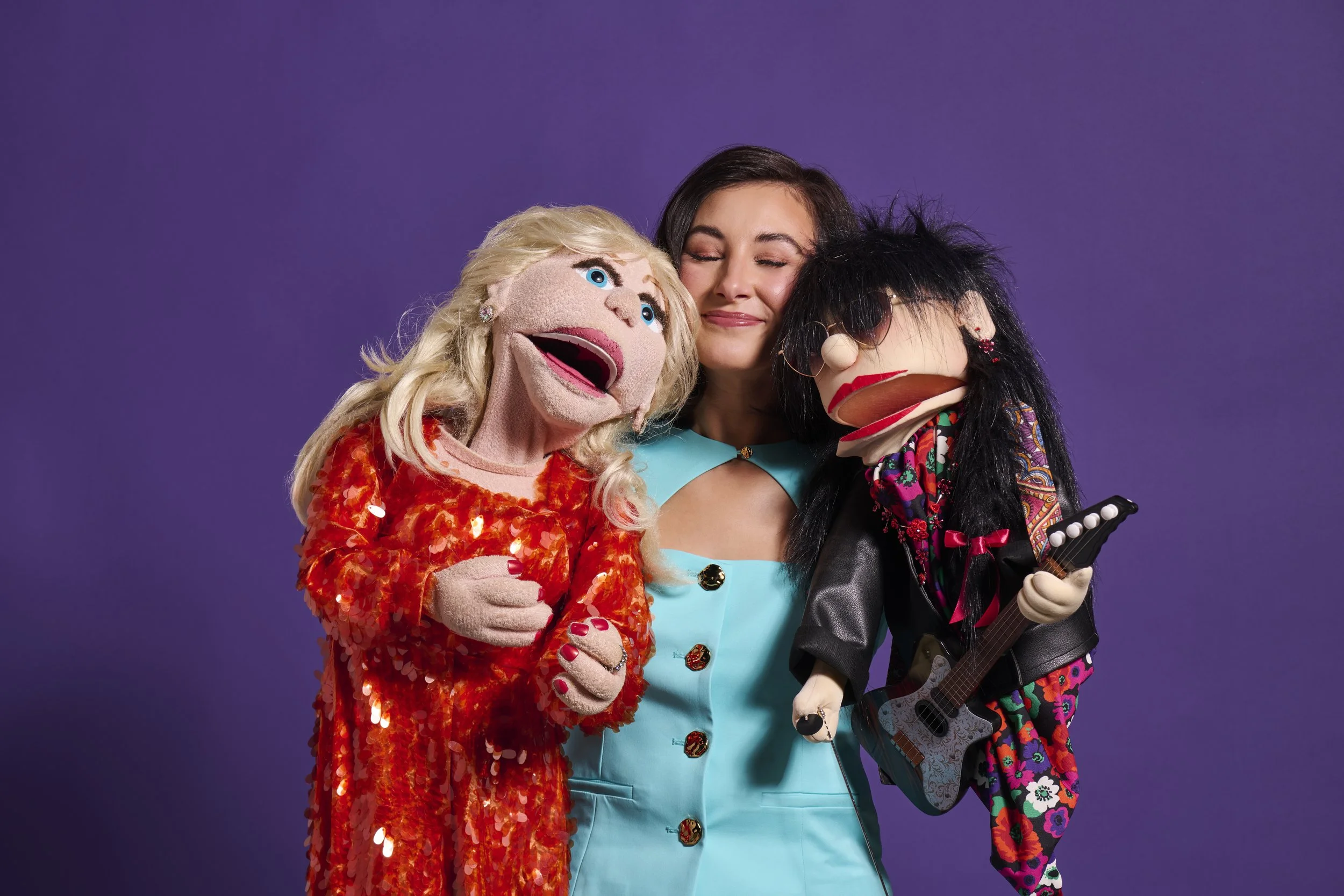 A woman hugging two puppets, one dressed in a red sequined outfit resembling a singer and the other in rock attire with a guitar puppet, against a purple background.