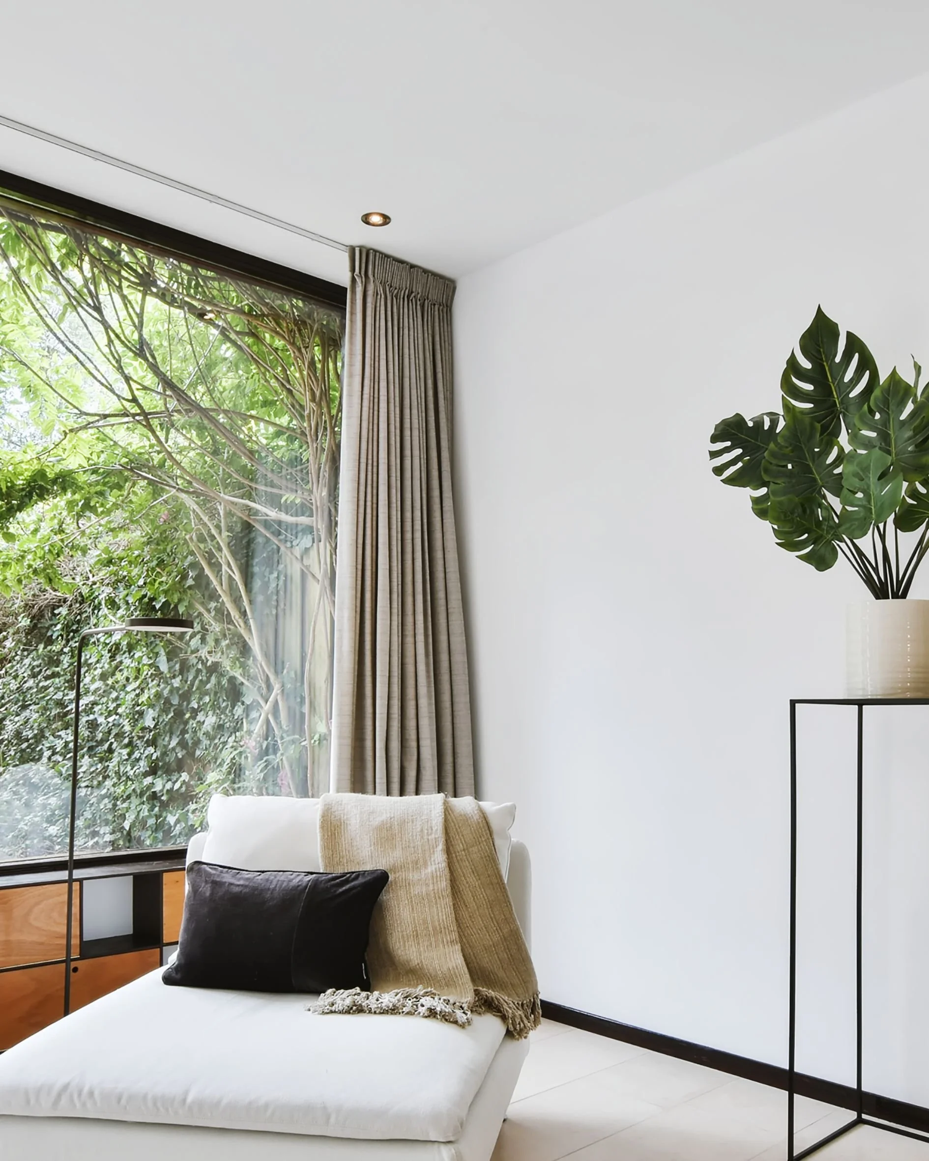 Modern living room with a large window, beige curtains, a white lounge chair with a black pillow and a beige blanket, and a tall green potted plant on a black metal stand.