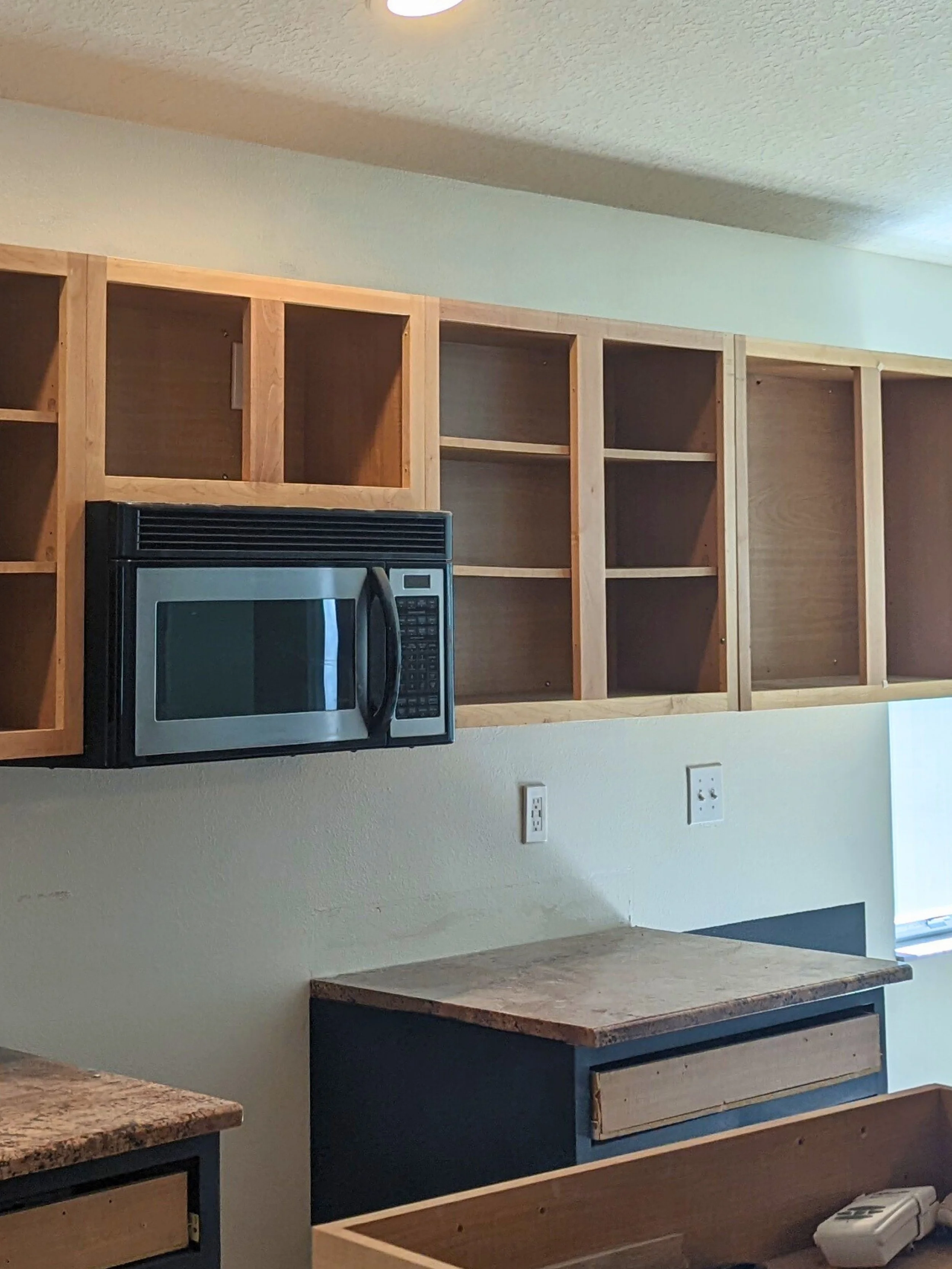 Unfinished kitchen with open wooden cabinets, a microwave, and granite countertops.