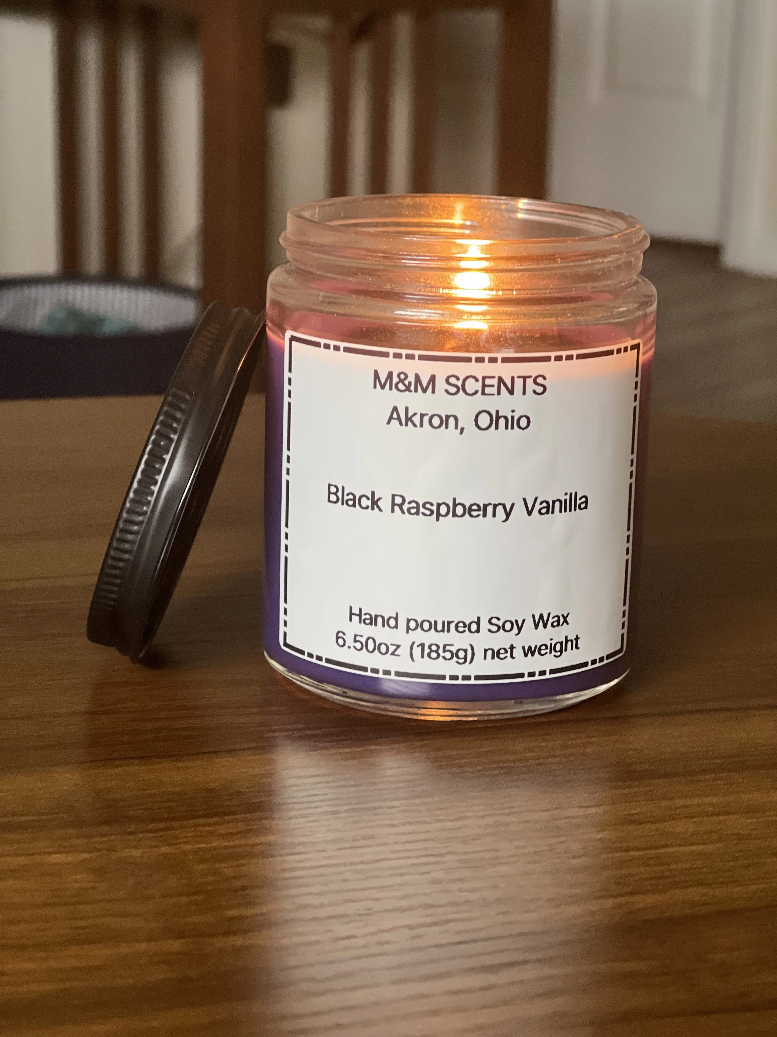 A lit candle in a glass jar with a black lid leaning against it. The candle label reads 'M&M SCENTS Akron, Ohio Black Raspberry Vanilla Hand poured Soy Wax 6.5oz (185g) net weight.' The candle is on a wooden table.
