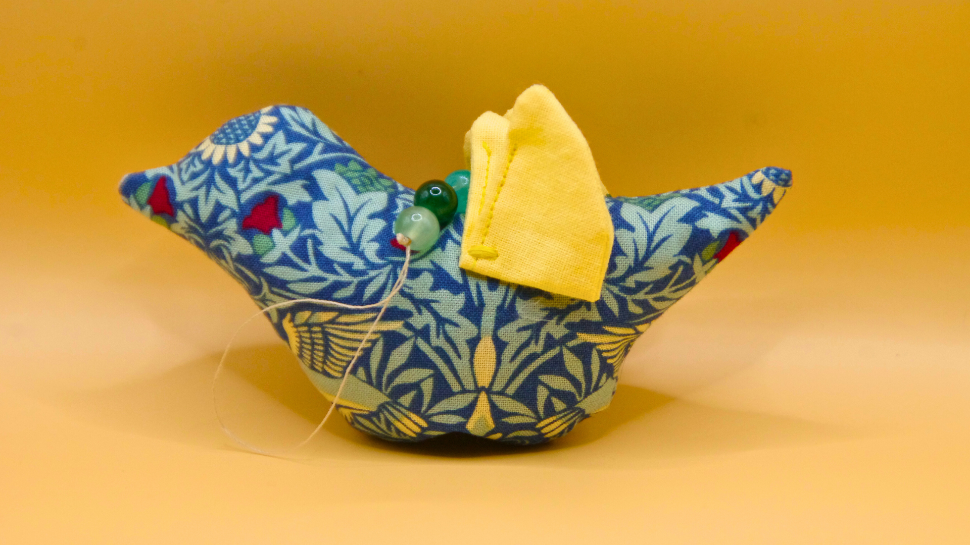 Fabric bird with blue and yellow floral pattern, yellow fabric wing, and beaded eye, set against a yellow background.