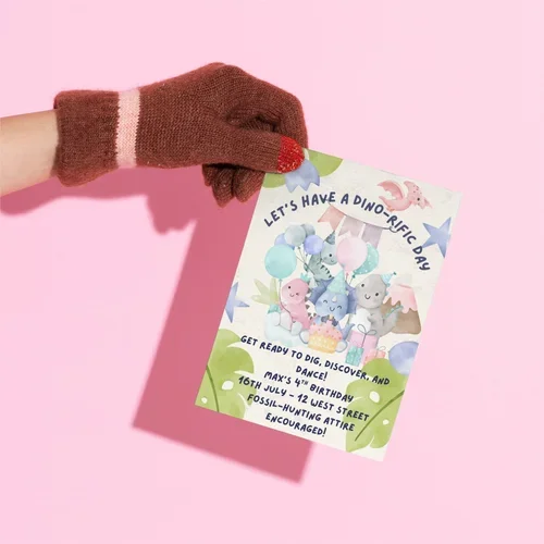A hand wearing a brown glove with pink and white stripes holding a colorful birthday invitation against a pink background. The invitation features illustrated dinosaurs and says, "Let's Have a Dino-rific Day" and provides details for a birthday party.