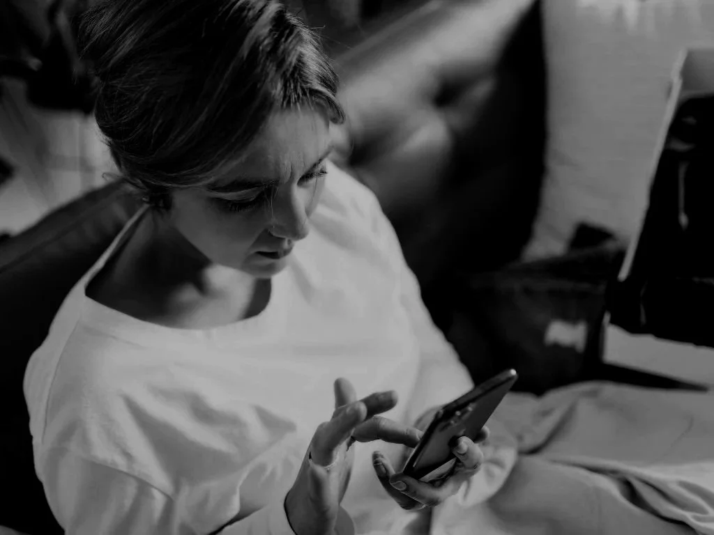 A woman sitting on a couch looking down and using a smartphone, wearing a white shirt with her short hair styled back, in a black and white photo.