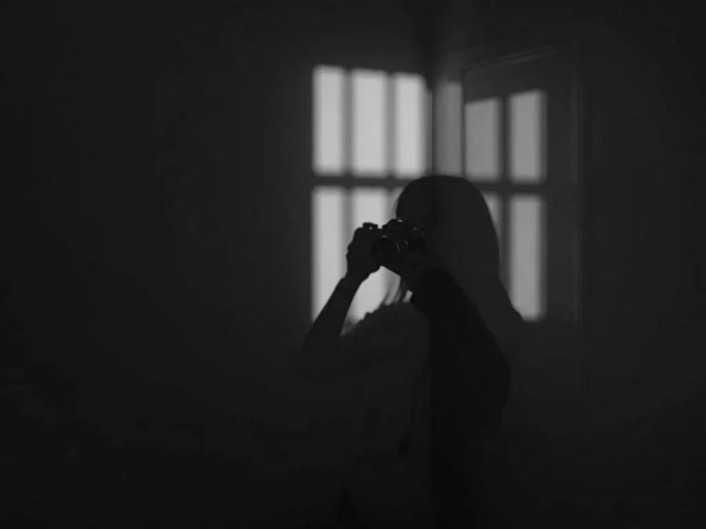 A person taking a photo with a camera, silhouette against a window with sunlight, in a dark room.