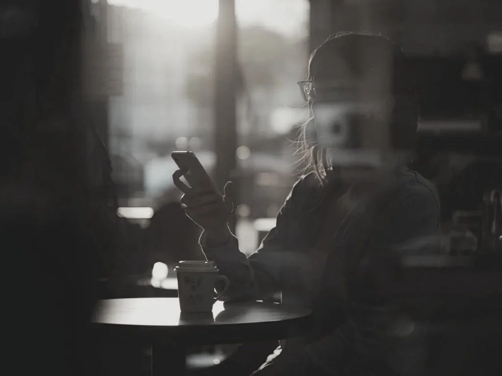 A woman with glasses sitting at a café table, looking at her phone, with a coffee mug in front of her, in a dimly lit setting.