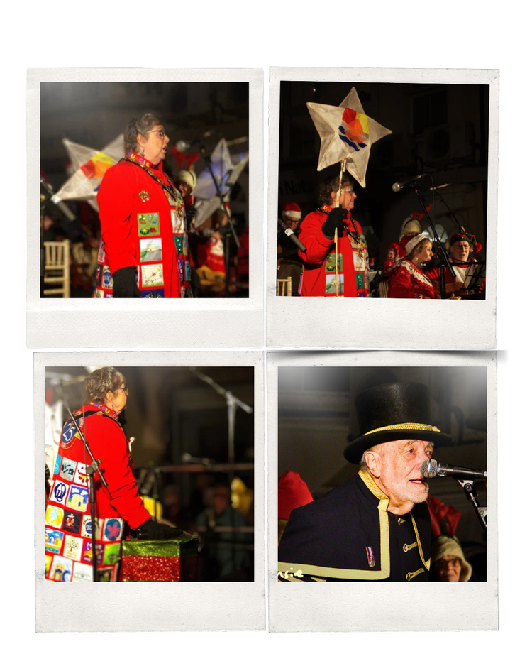 A Christmas-themed choir and band performing on stage. The performers are dressed in festive, colorful costumes, some with holiday hats. There are microphones, Christmas decorations, and a dark background indicating an indoor event.