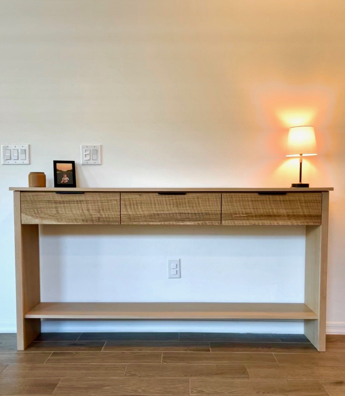 Wooden console table with three drawers, a small framed picture, a candle, and a lit table lamp on top, against a plain white wall with electrical outlets.