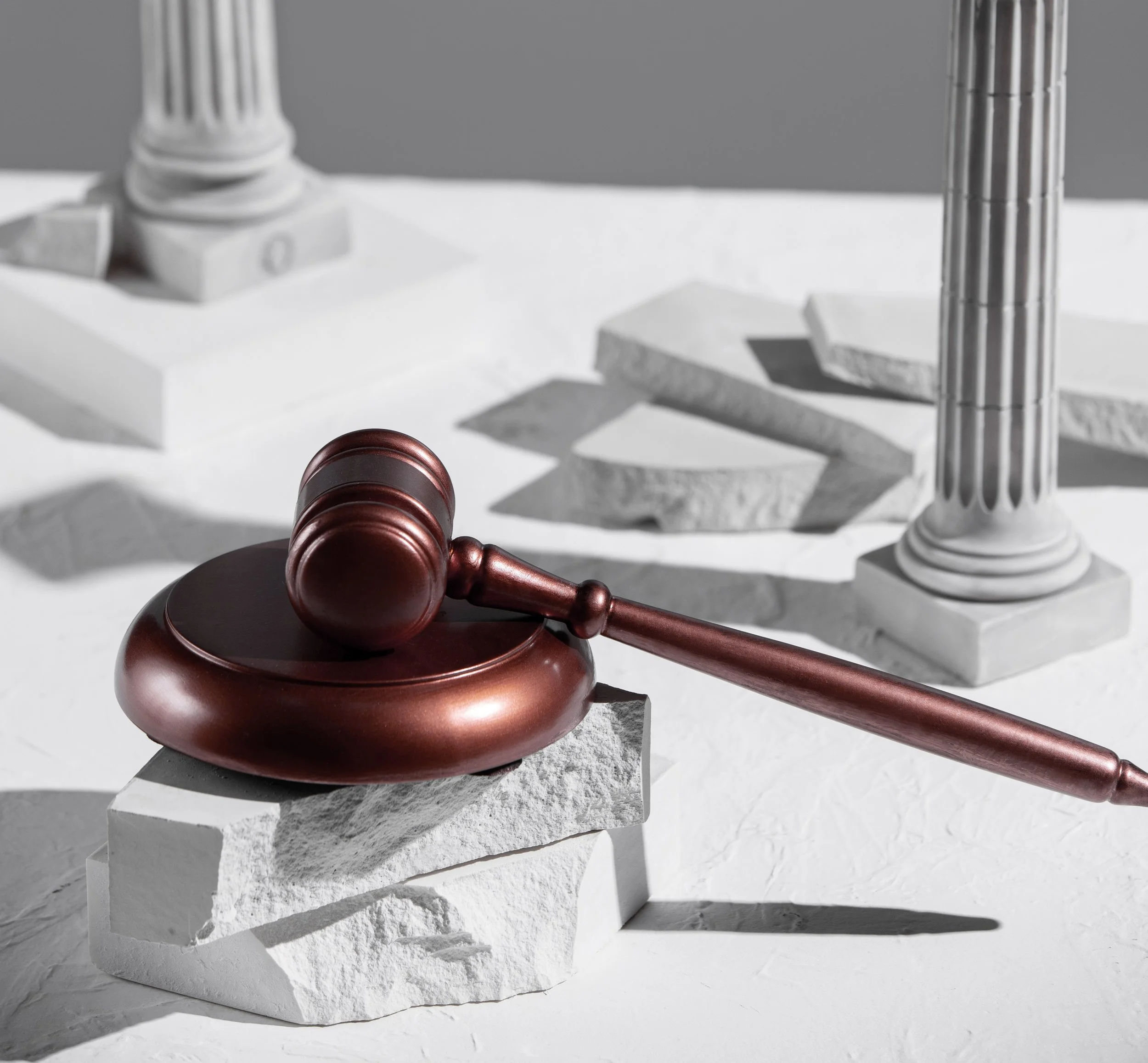 A judge's gavel resting on a round base, with white and grey broken columns and pedestals in the background, set on a white textured surface.